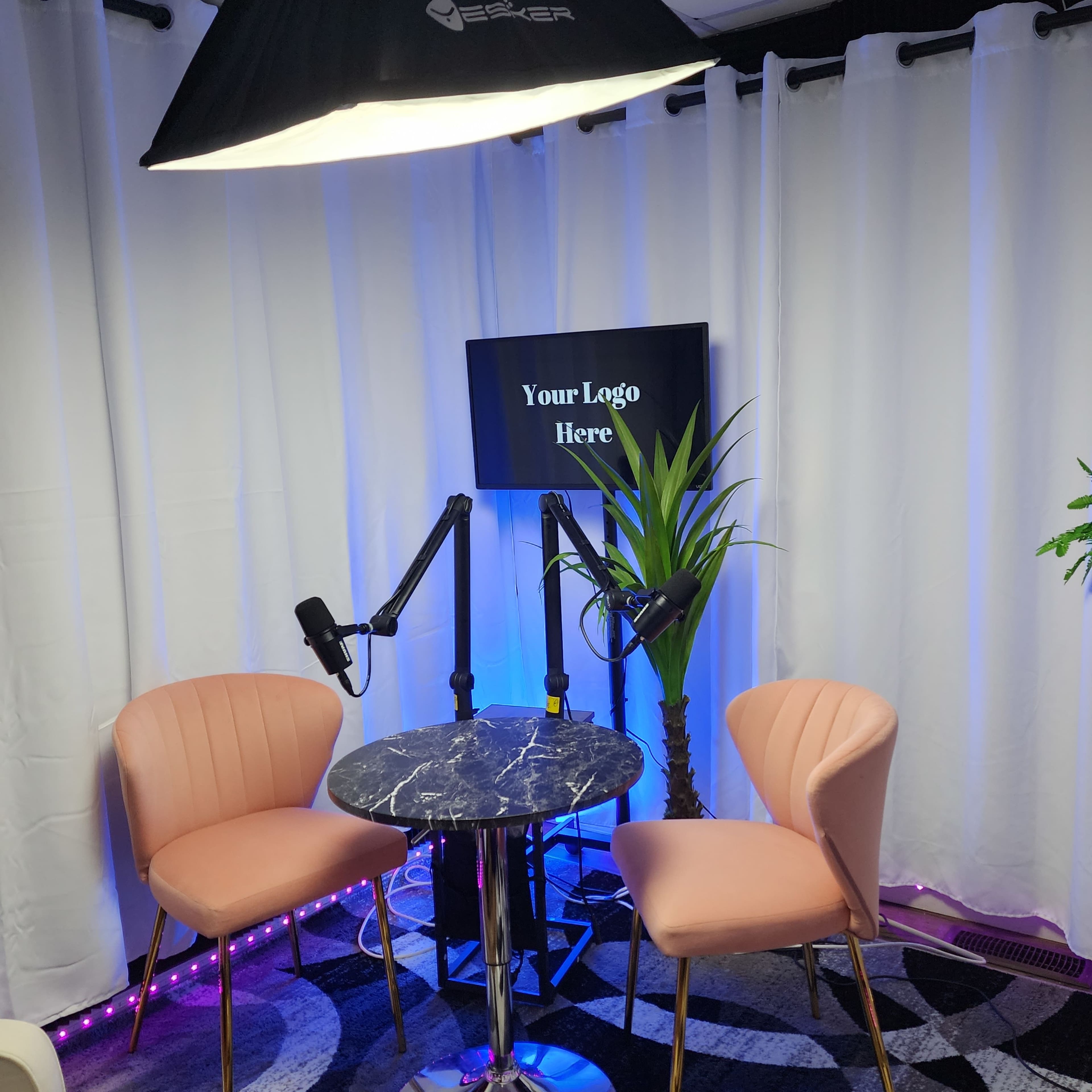 A small studio setup with two pink chairs, a circular marble table, and a screen displaying the text "Your Logo Here," surrounded by white curtains and decorative plants.