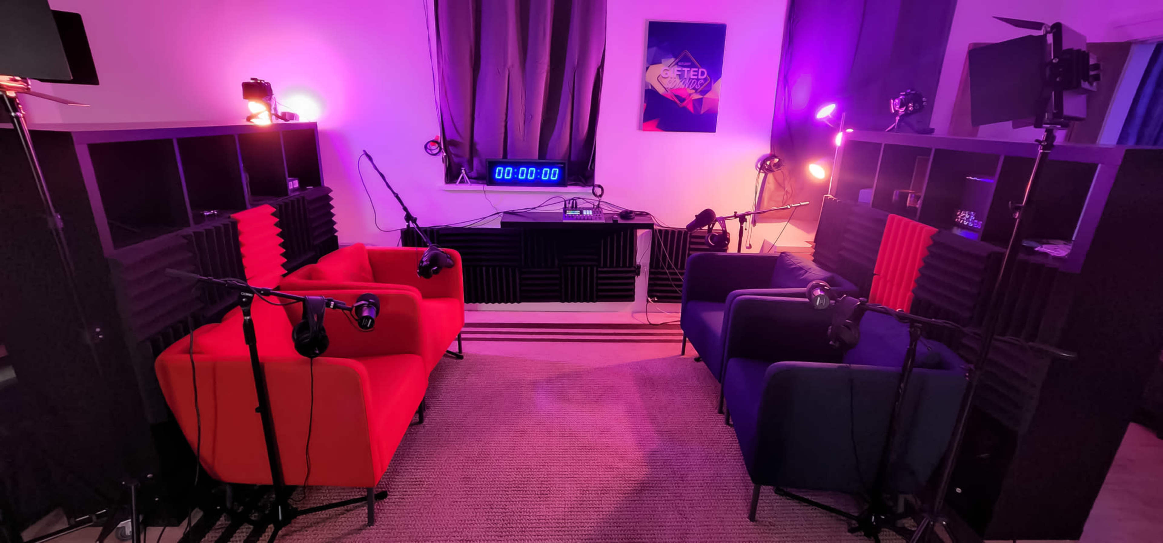 A recording studio setup with two colored armchairs, microphones, and a digital timer in the center, surrounded by lighting and soundproofing panels.