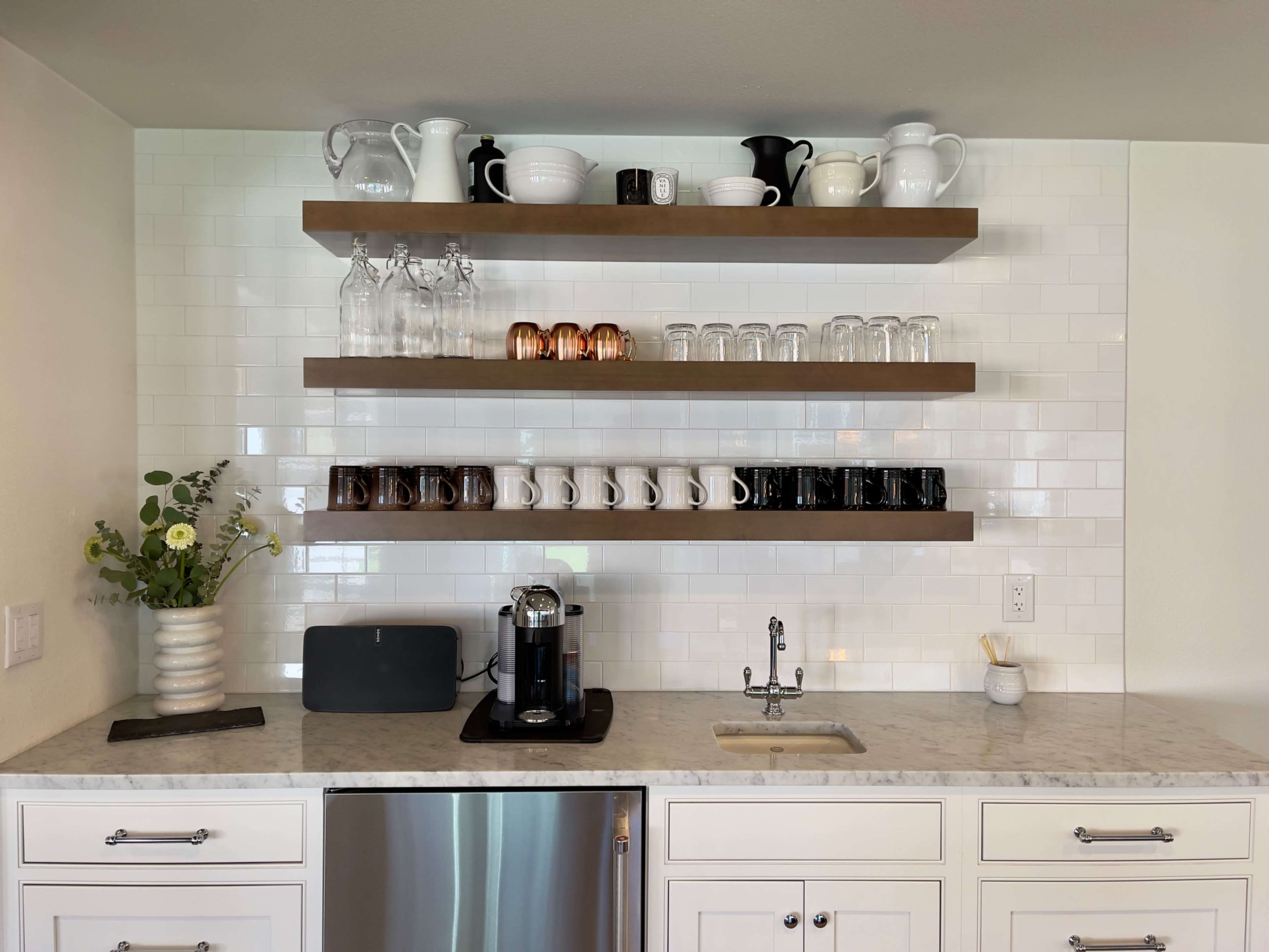 A modern kitchen features open shelves with various ceramic and glass items arranged above a countertop with a coffee maker and a sink.