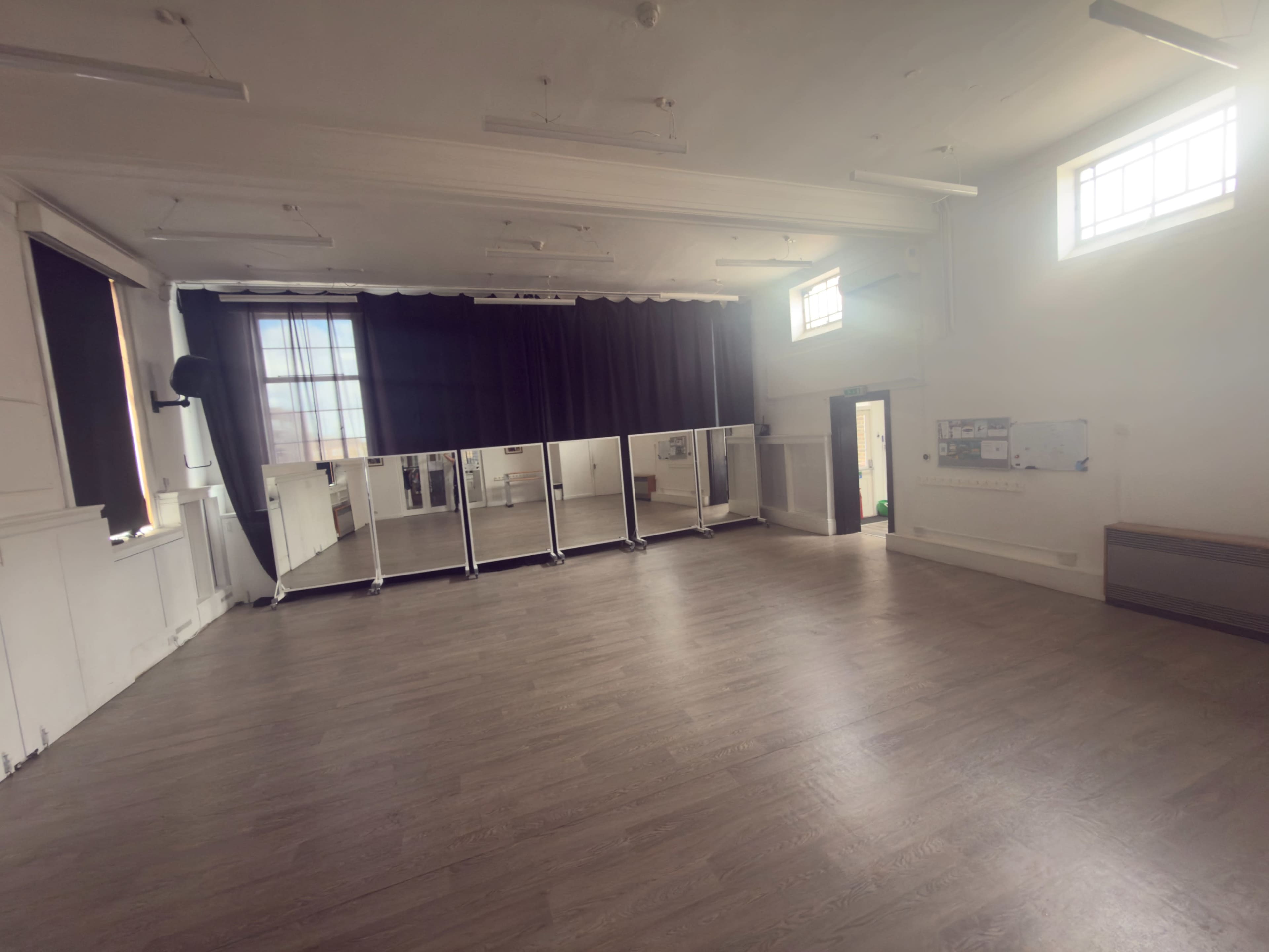 The image shows a spacious room with wooden flooring, large mirrors lined against one wall, and windows allowing natural light to enter.
