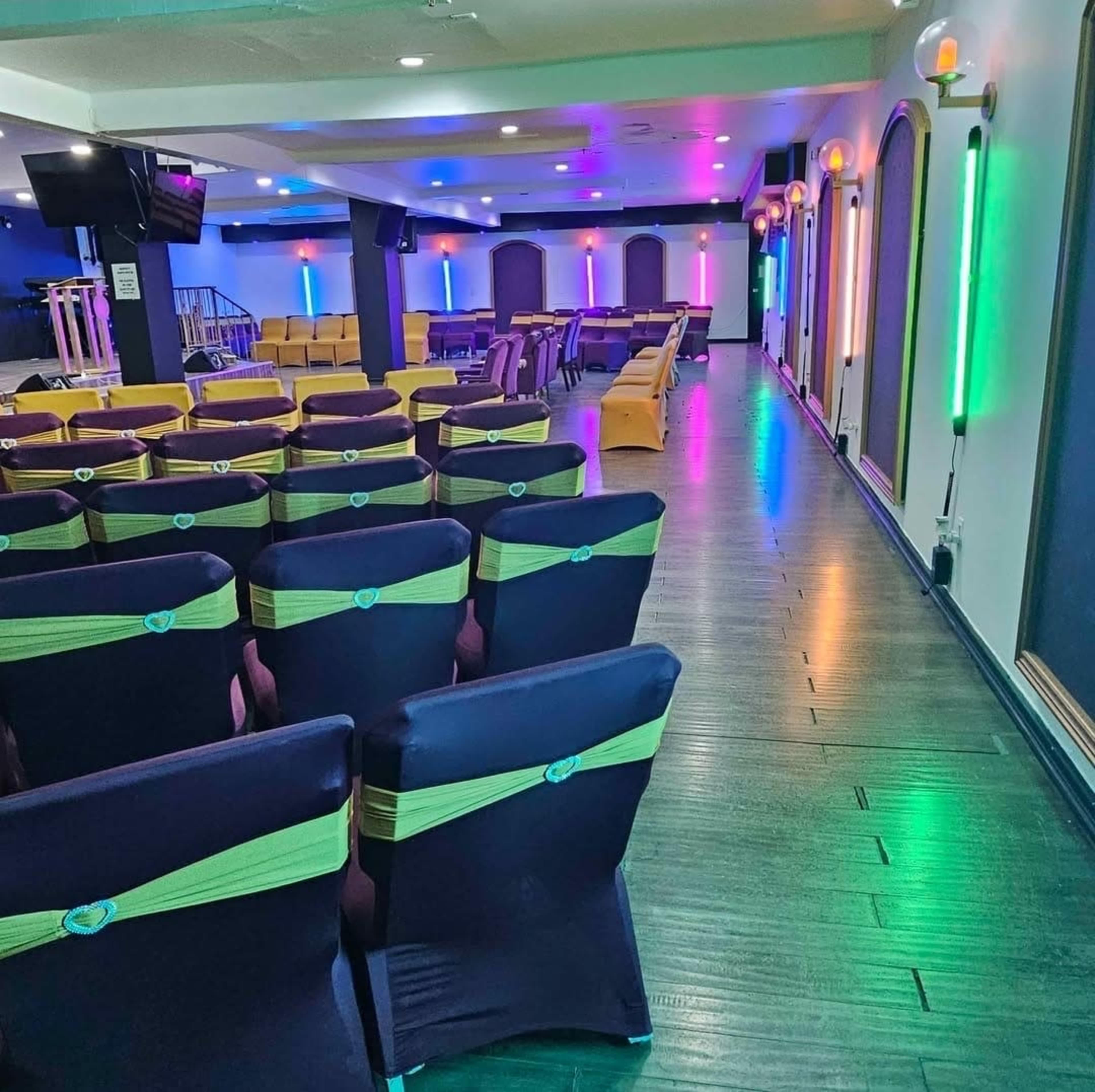 The image shows a brightly lit event space featuring rows of chairs with green sashes, colorful accent lighting, and a stage area in the background.