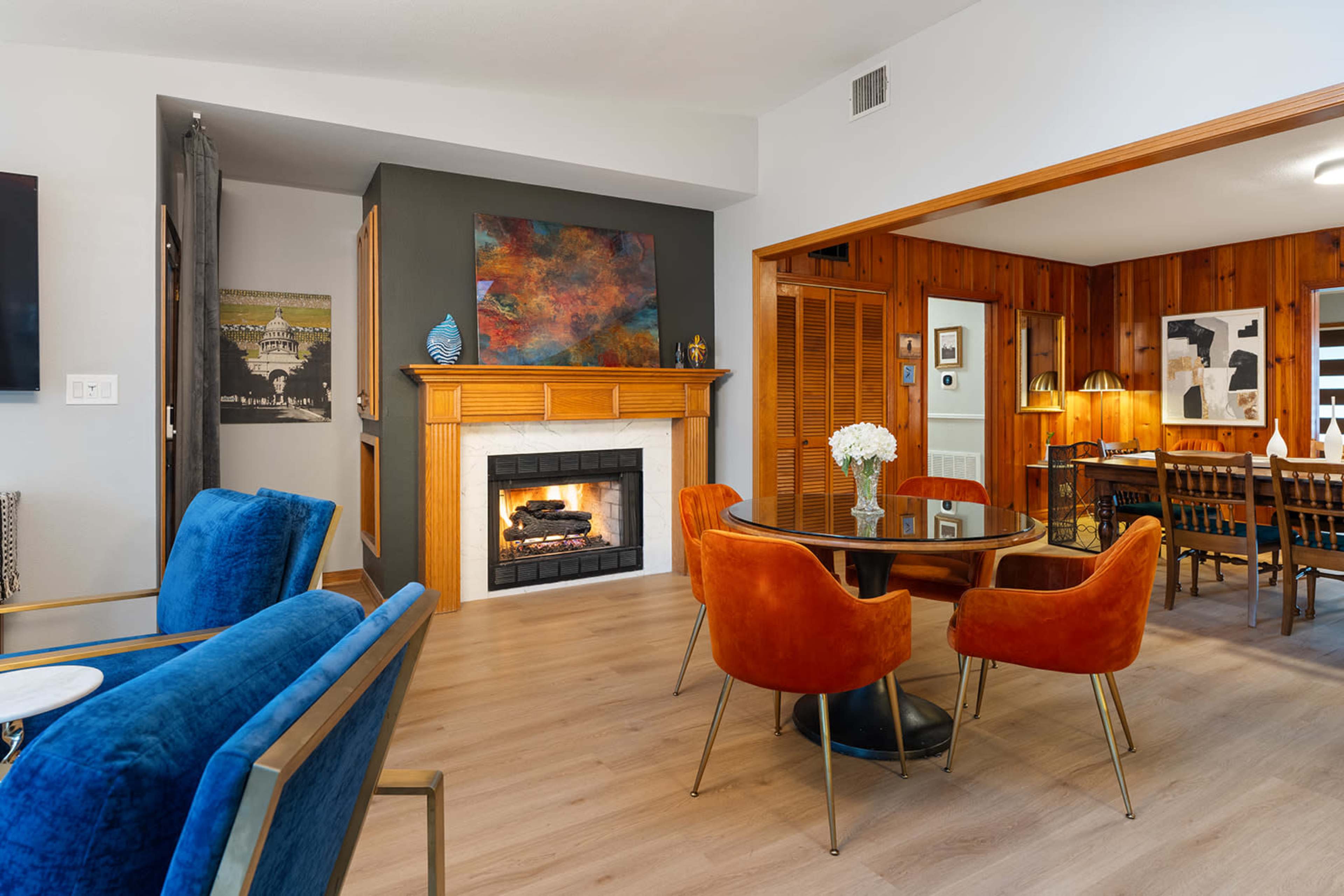 The image depicts a modern living space featuring a fireplace, a round dining table with orange chairs, and wood-paneled walls.