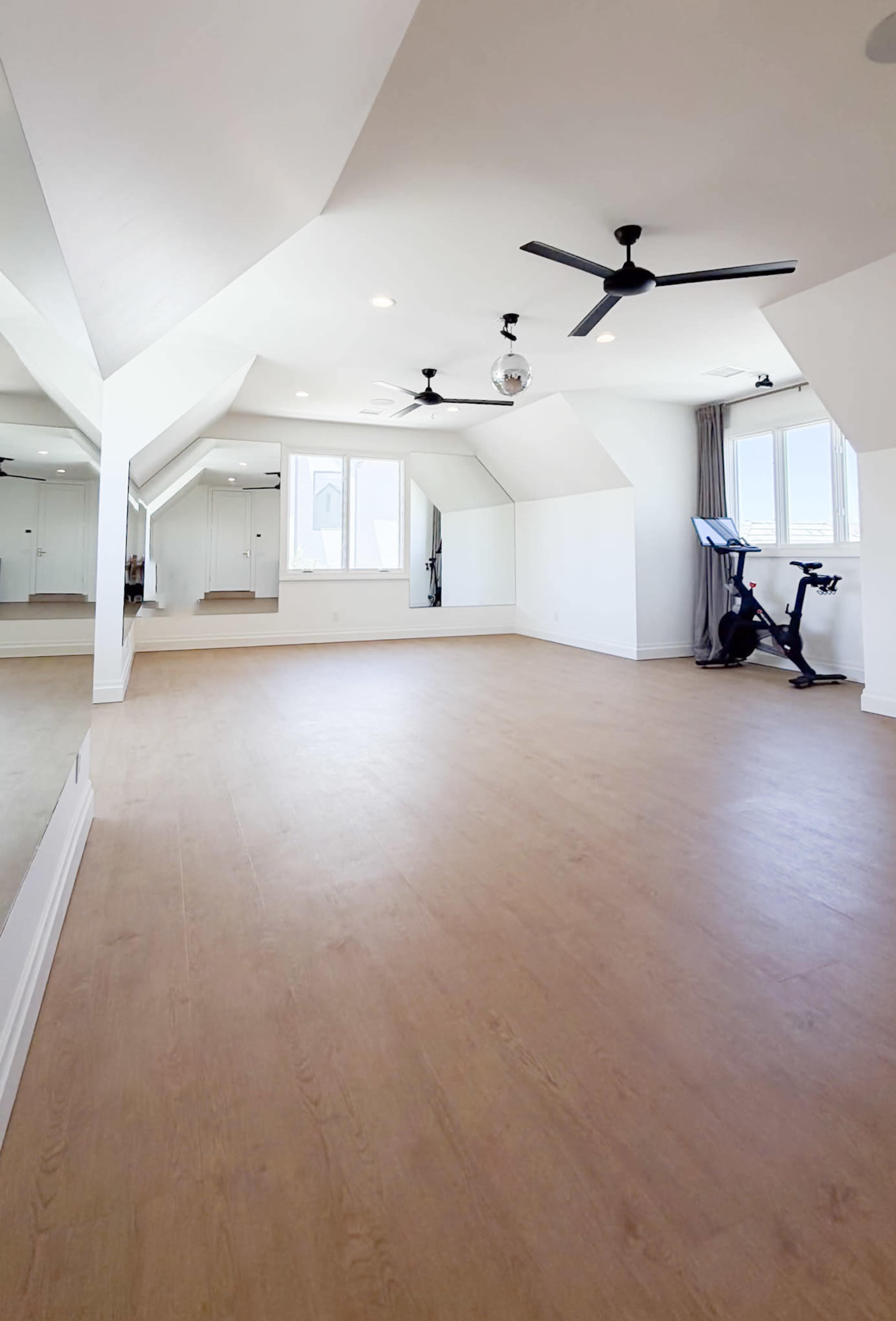The image shows a spacious room with wooden flooring, large windows, a stationary bike, and mirrors along one wall.