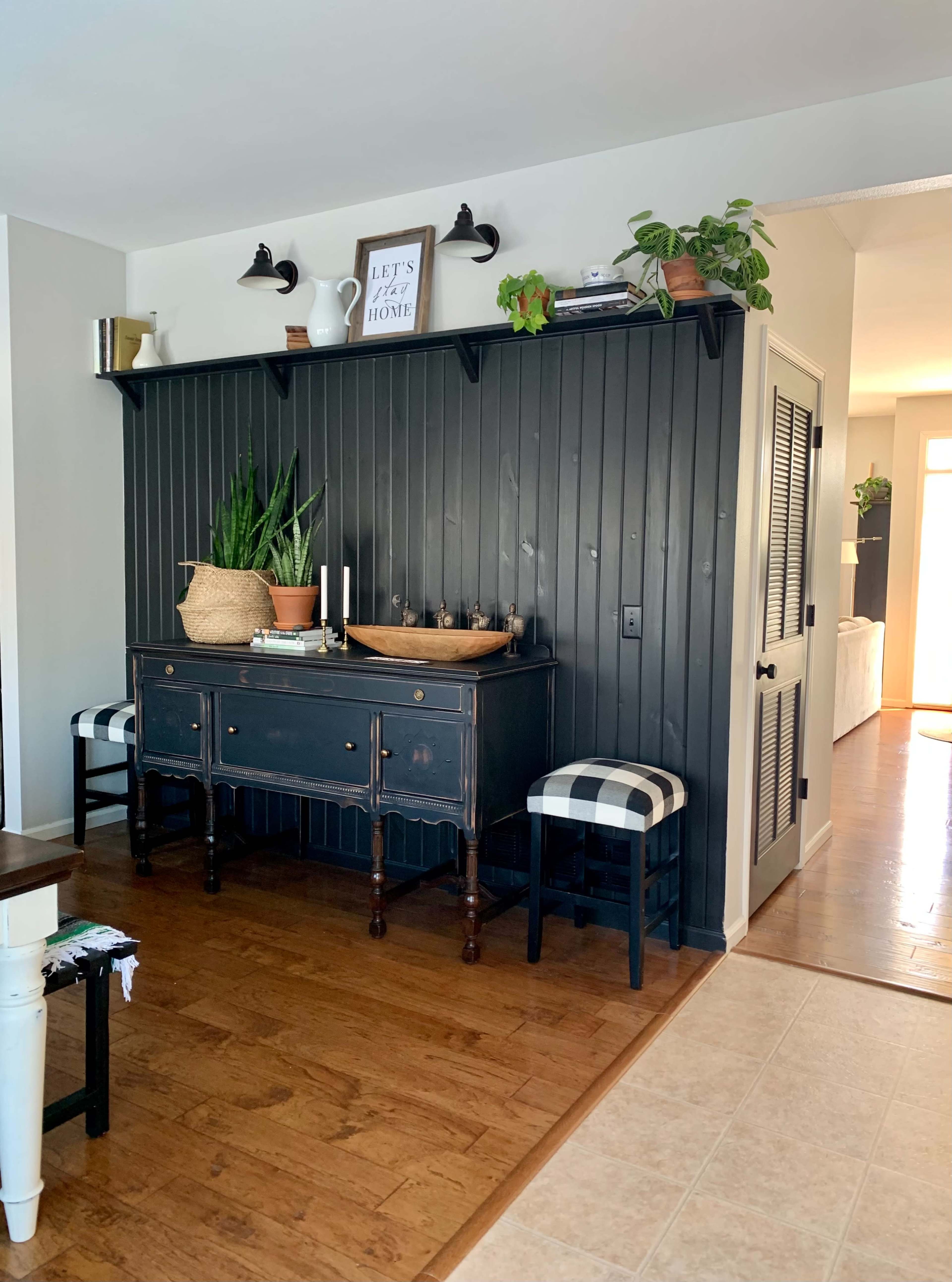 A dark wooden sideboard is placed against a paneled wall, accompanied by decorative items and plants, with two checkered stools nearby.