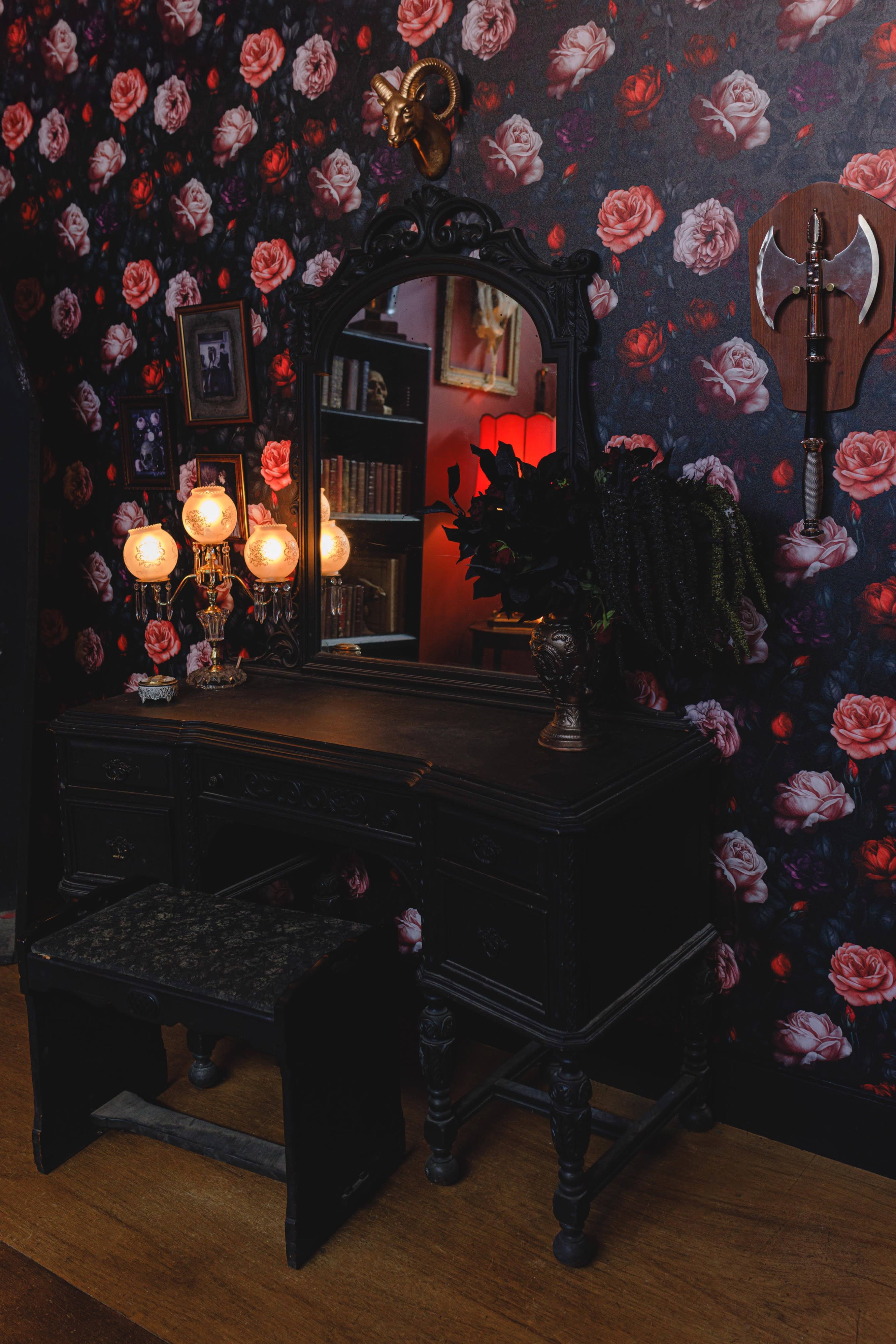 A black vanity dresser with a large mirror is positioned against a floral wallpaper featuring red roses, accompanied by a decorative axe on the wall and illuminated by two lamps.