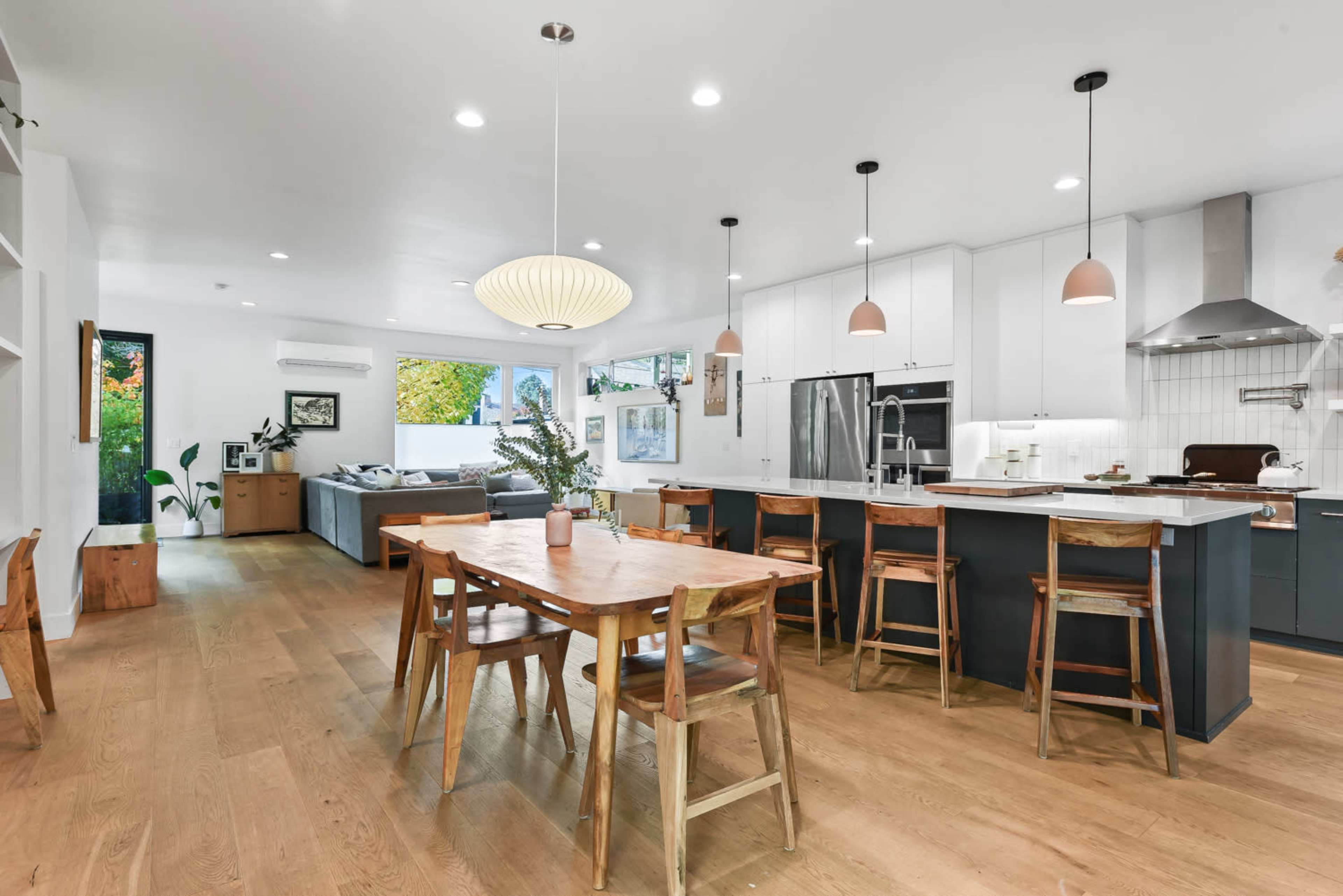 A modern kitchen and dining area features a wooden table surrounded by chairs, sleek cabinetry, and large windows allowing natural light to fill the space.