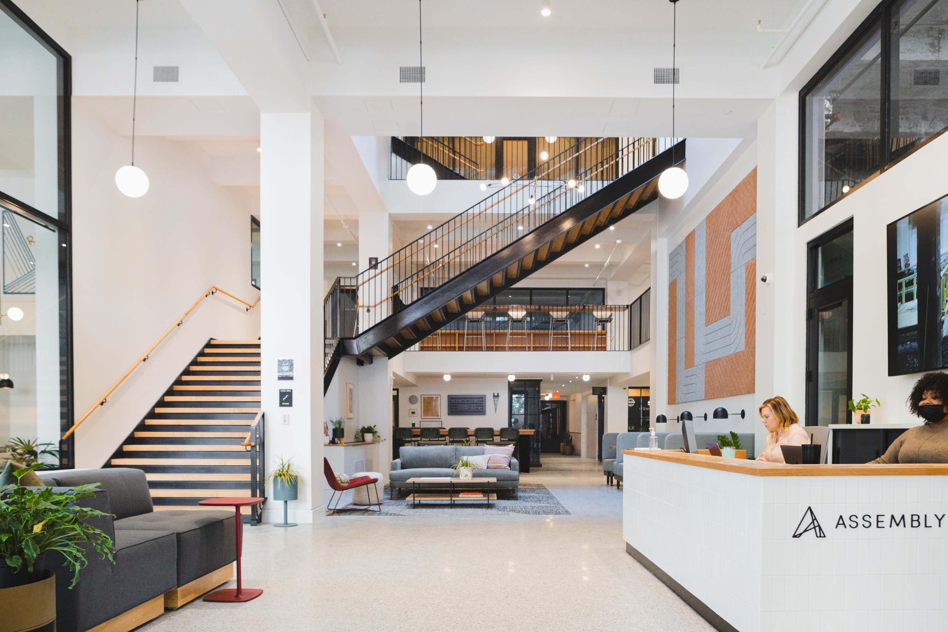 The image shows a modern lobby with a reception desk, seating areas, and a staircase leading to an upper level, filled with natural light and greenery.