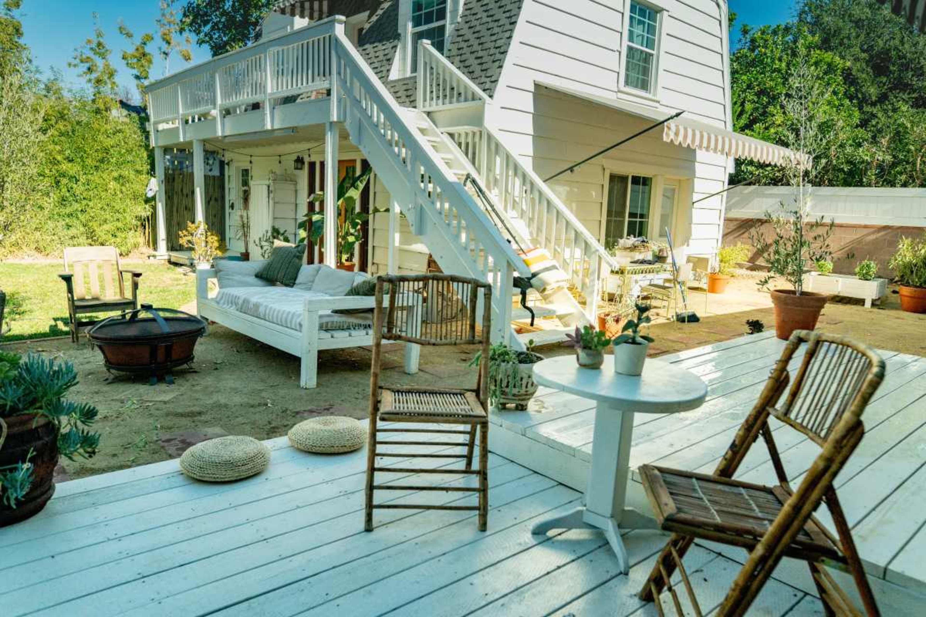 The image features a backyard scene with a two-story white house, wooden stairs, patio furniture, and a grassy area surrounded by potted plants.