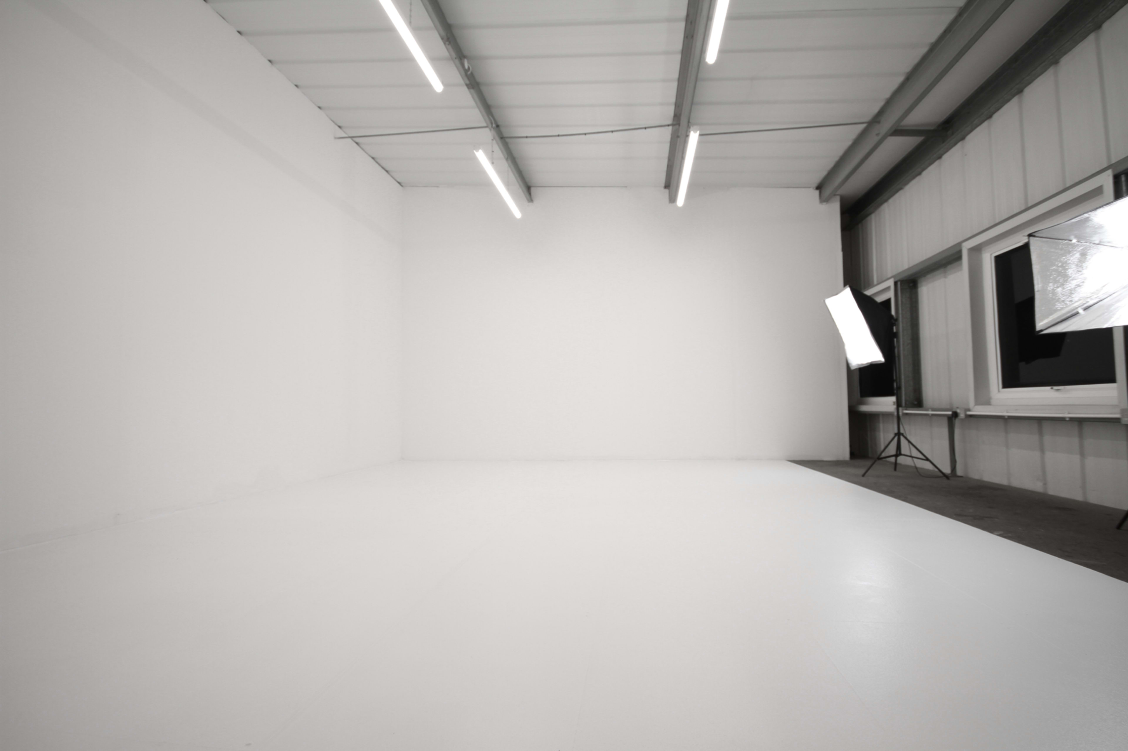 The image depicts a spacious, empty studio with white walls and a smooth floor, illuminated by overhead lighting and softbox lights positioned in the corner.