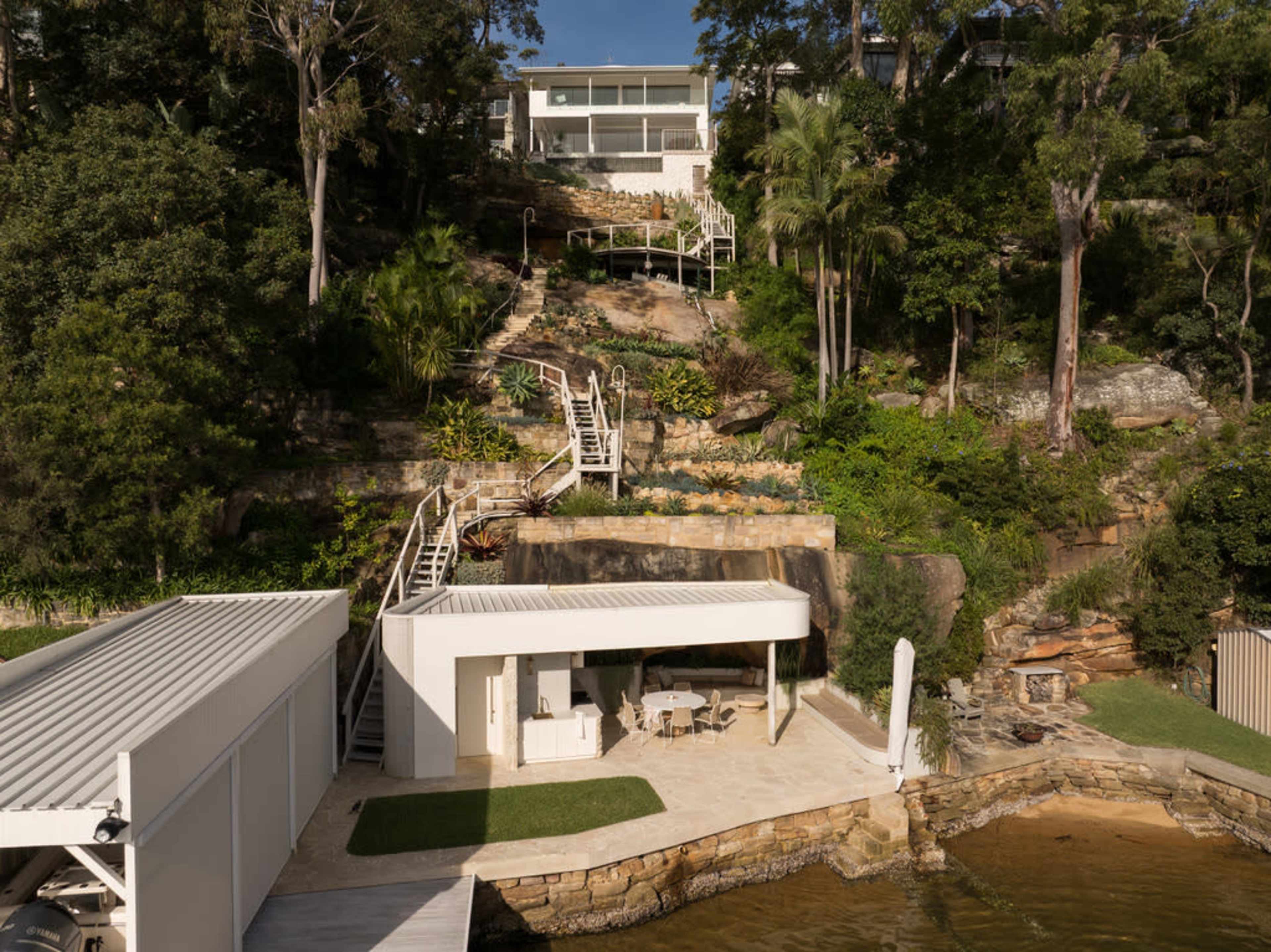 The image shows a modern house situated on a hillside with multiple staircases leading down to a waterfront area.