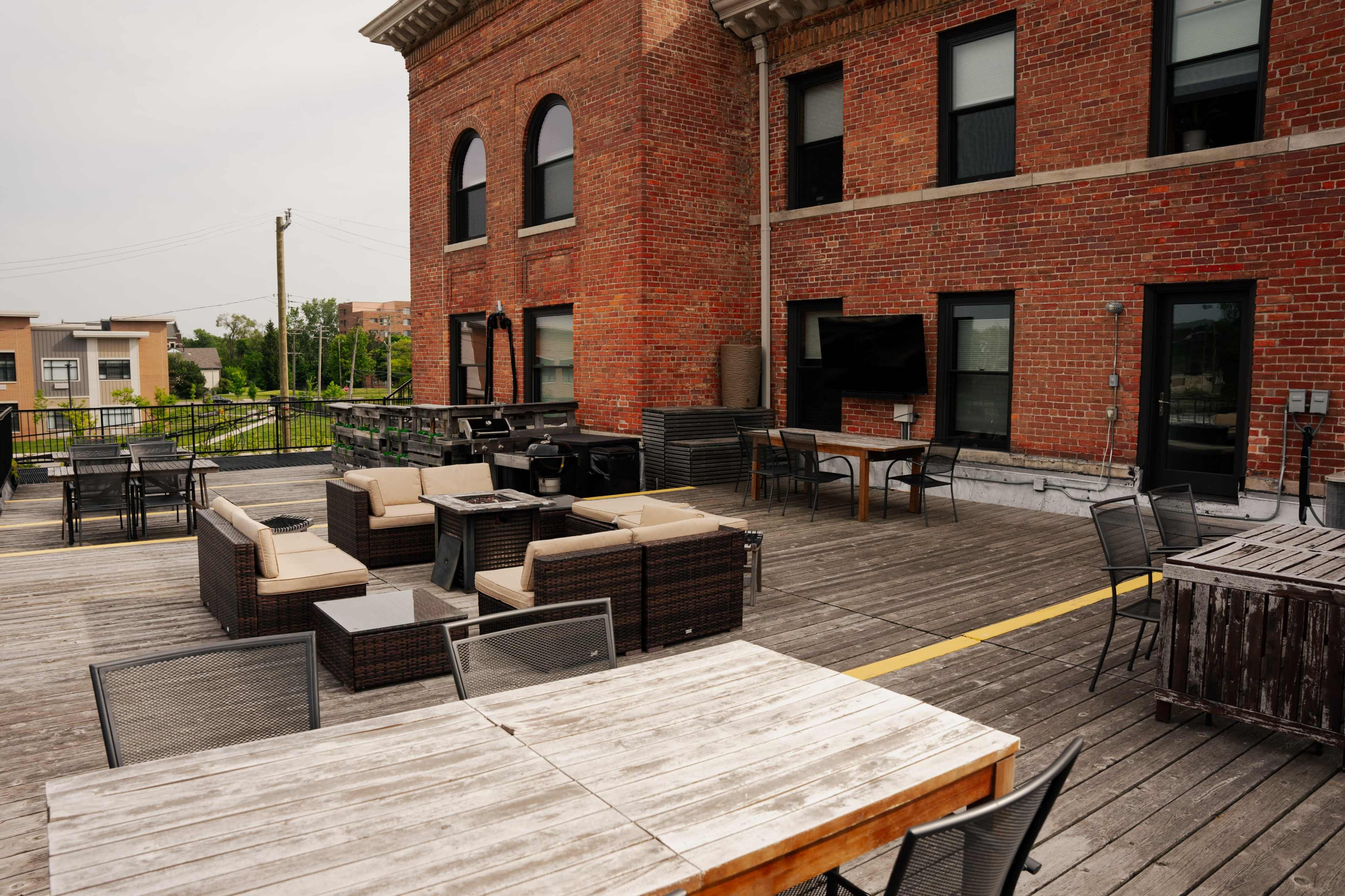 A spacious outdoor patio area with wicker furniture, wooden tables, and a large brick building in the background.
