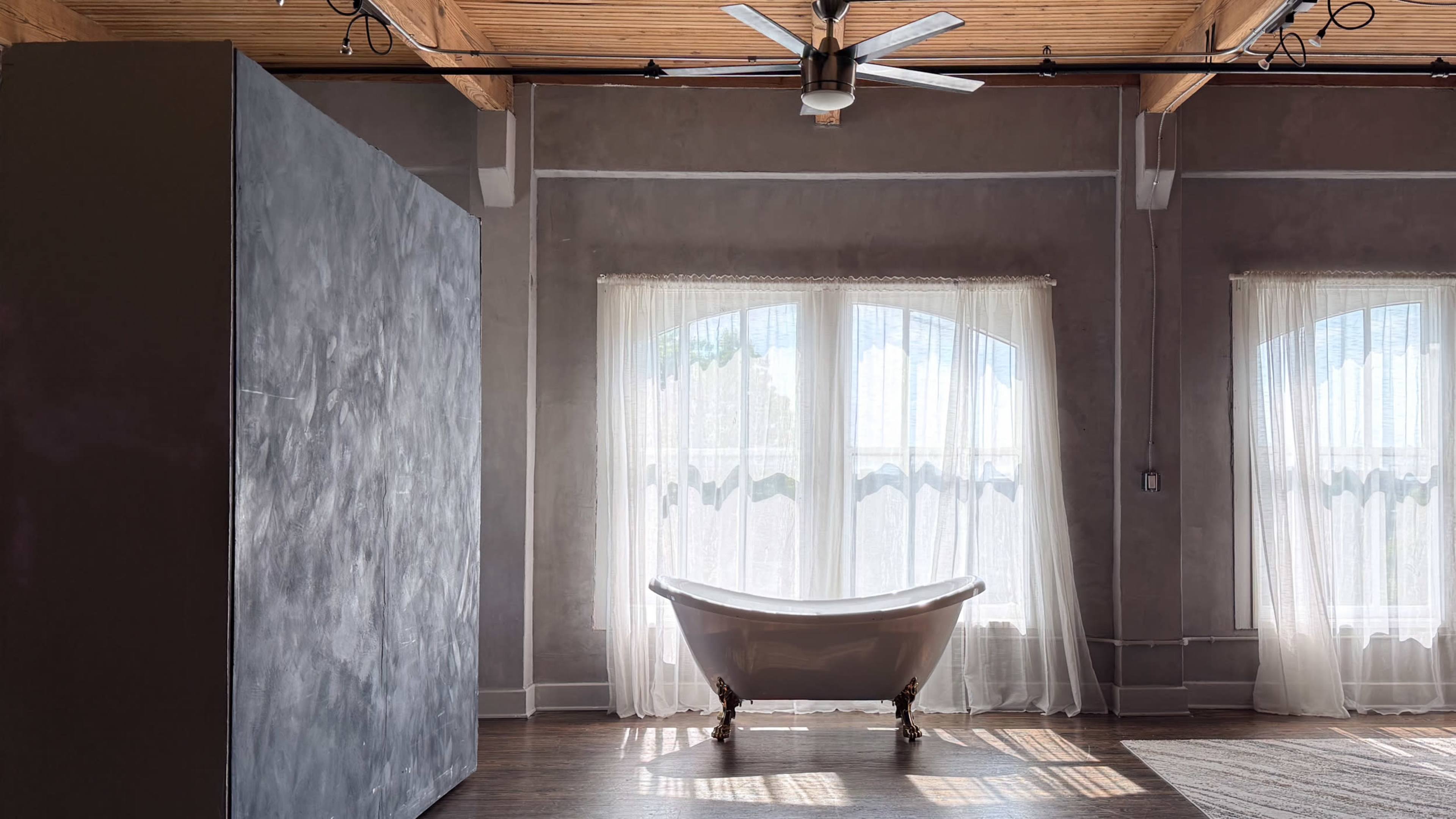 The image shows a bright room with large windows, featuring a vintage freestanding bathtub and high wooden ceilings.