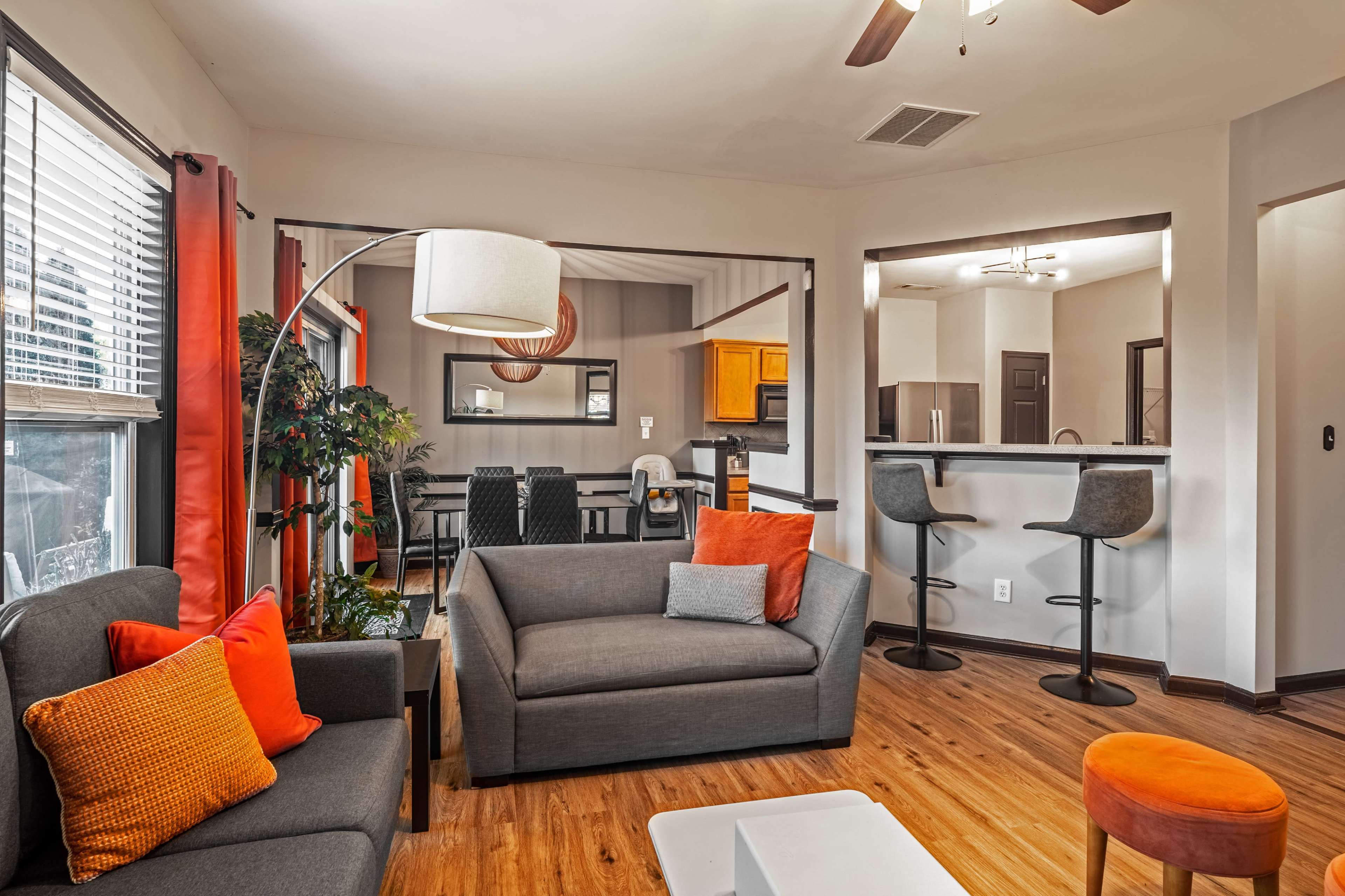 A living room area with a gray sofa, an orange accent chair, a floor lamp, and a view into a dining area with a kitchen in the background.