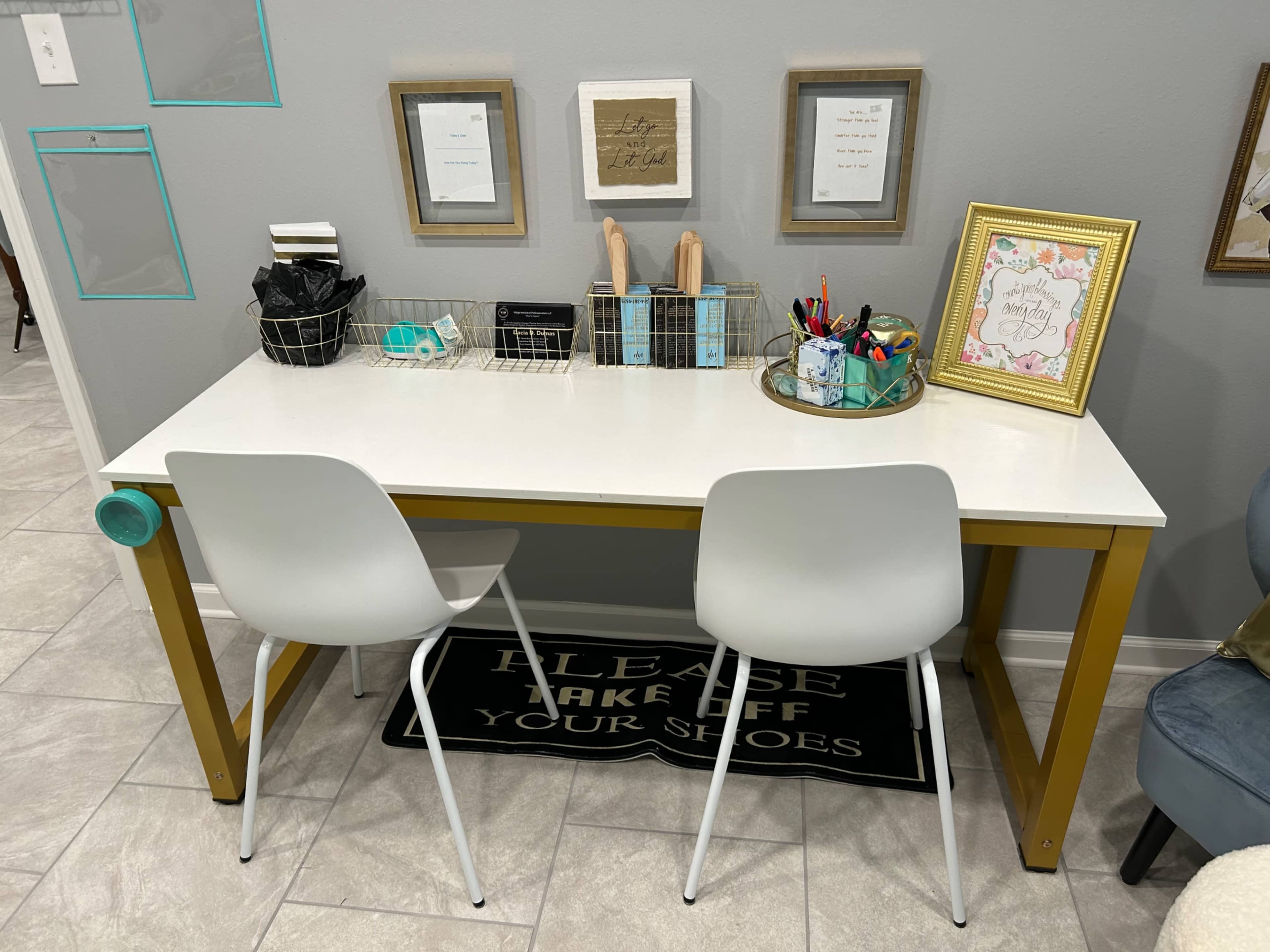 A modern desk with gold legs is adorned with various stationery items and framed pictures, accompanied by two white chairs and a "Please Take Off Your Shoes" rug.