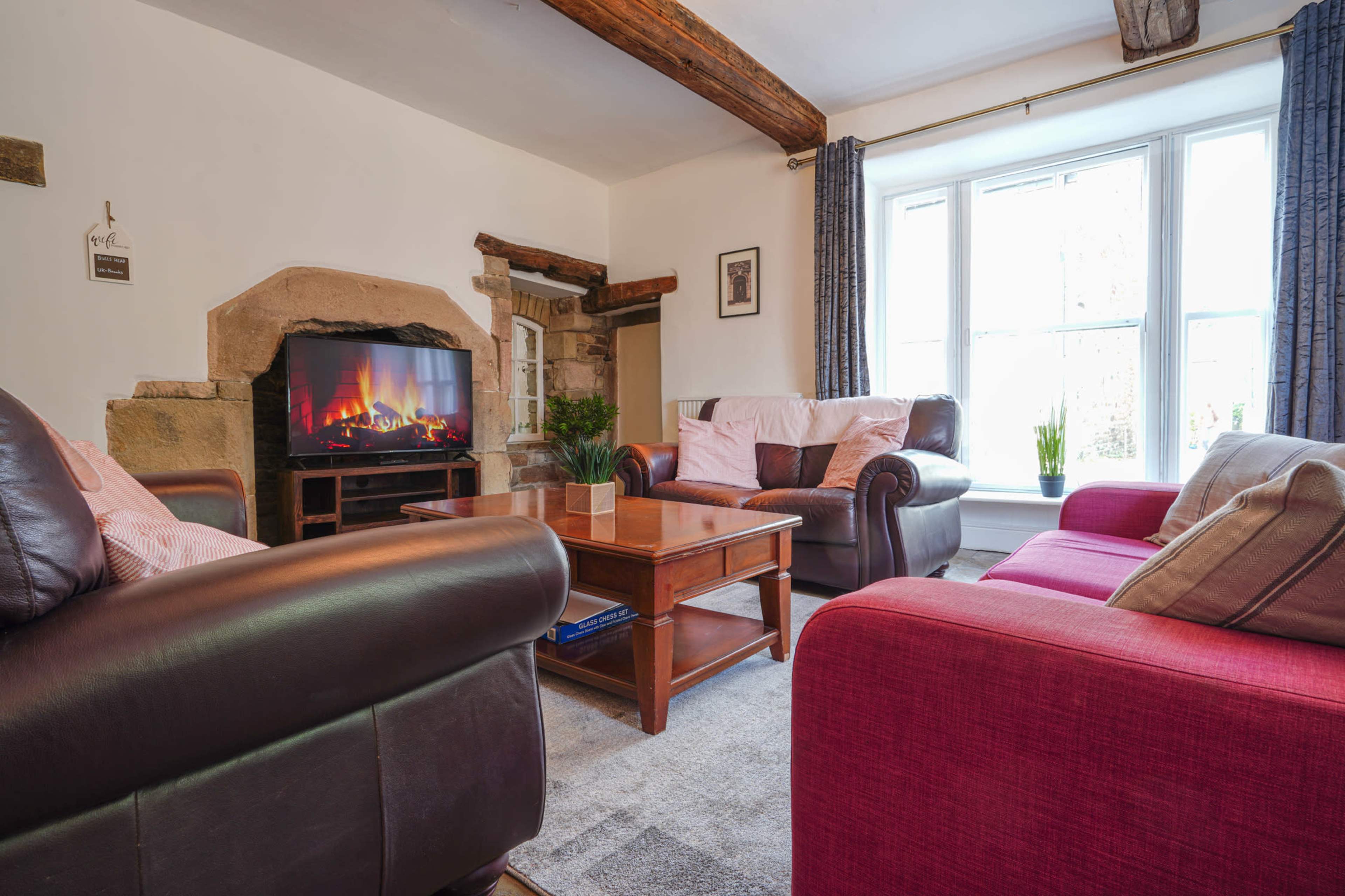 The image shows a cozy living room with leather sofas, a wooden coffee table, and a television displaying a fireplace, situated by a window with natural light.
