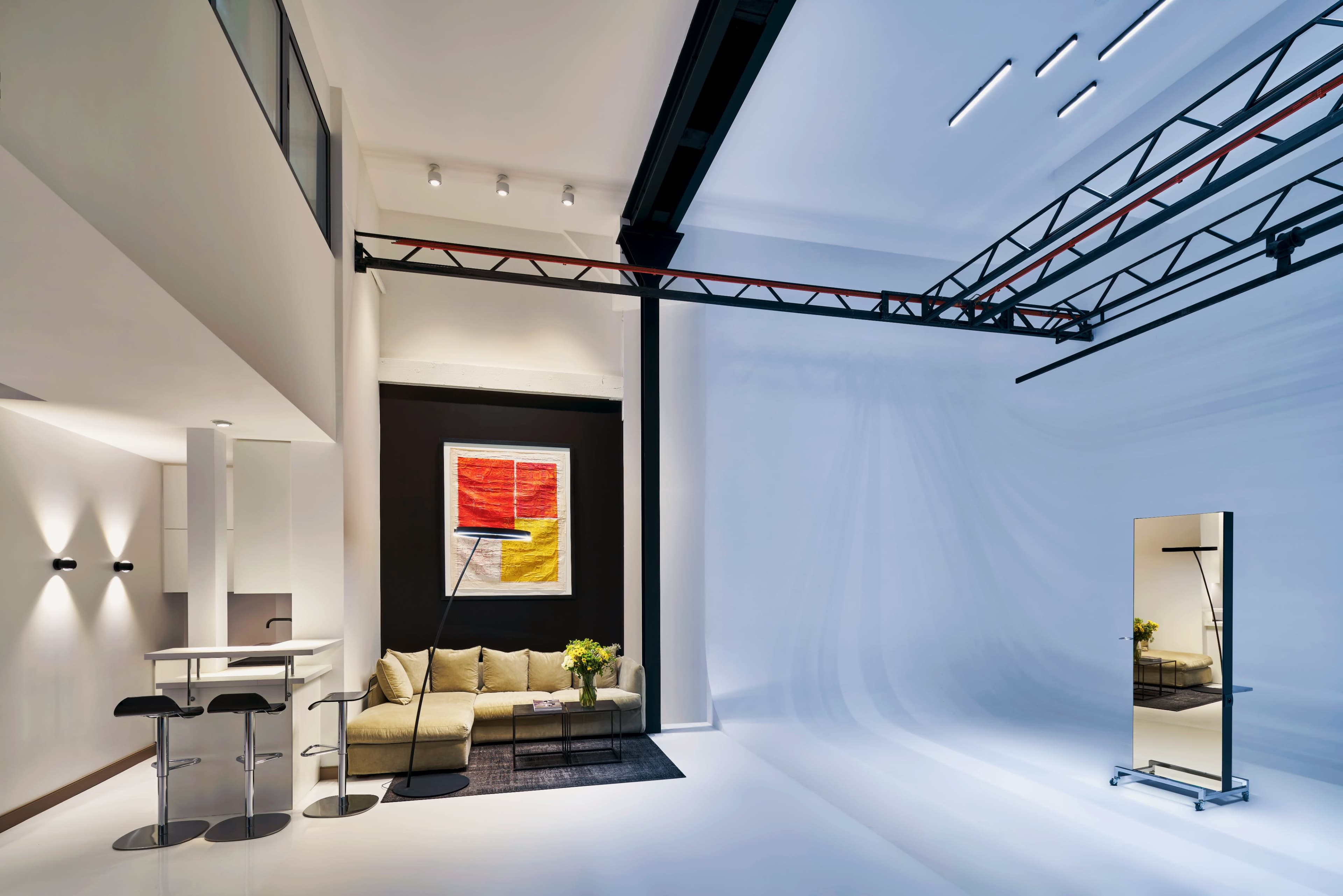 The image shows a modern, minimally furnished living space with a sofa, a mirror, and a large piece of abstract artwork against a stark white background.