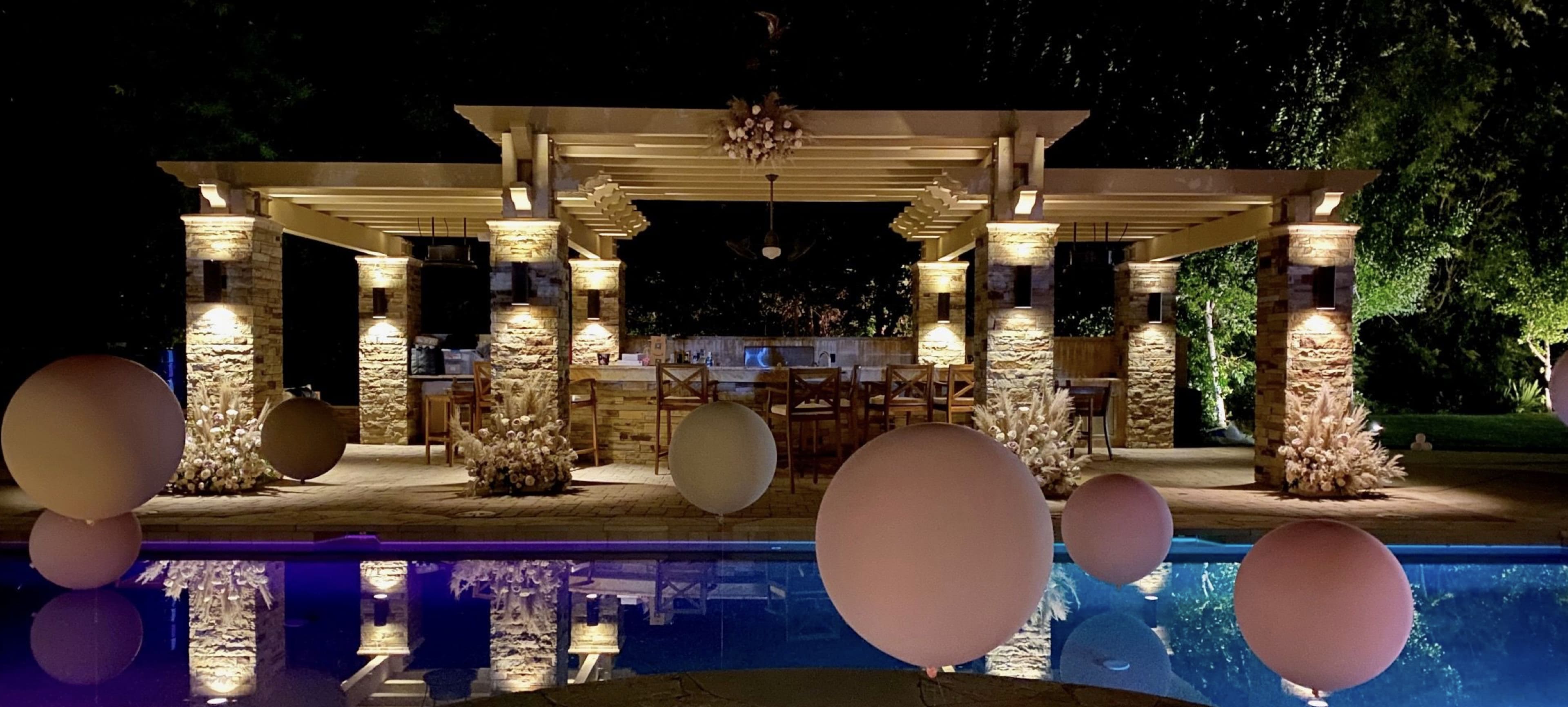 A illuminated patio area with a stone structure featuring columns, surrounded by large decorative spheres and reflecting in a pool.