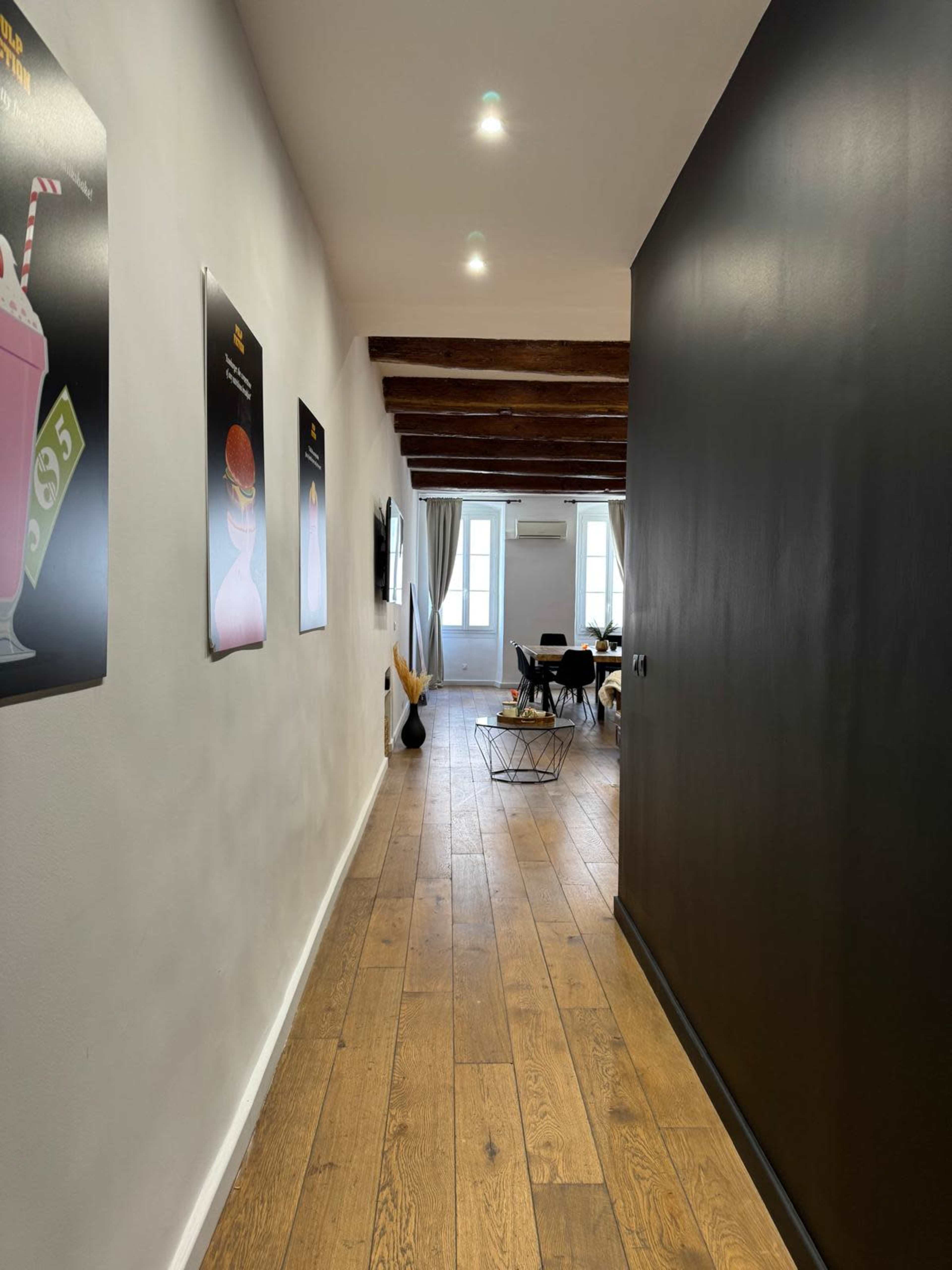 The image shows a hallway with wooden flooring, featuring black and white walls, and posters on the left side leading to a room with large windows.