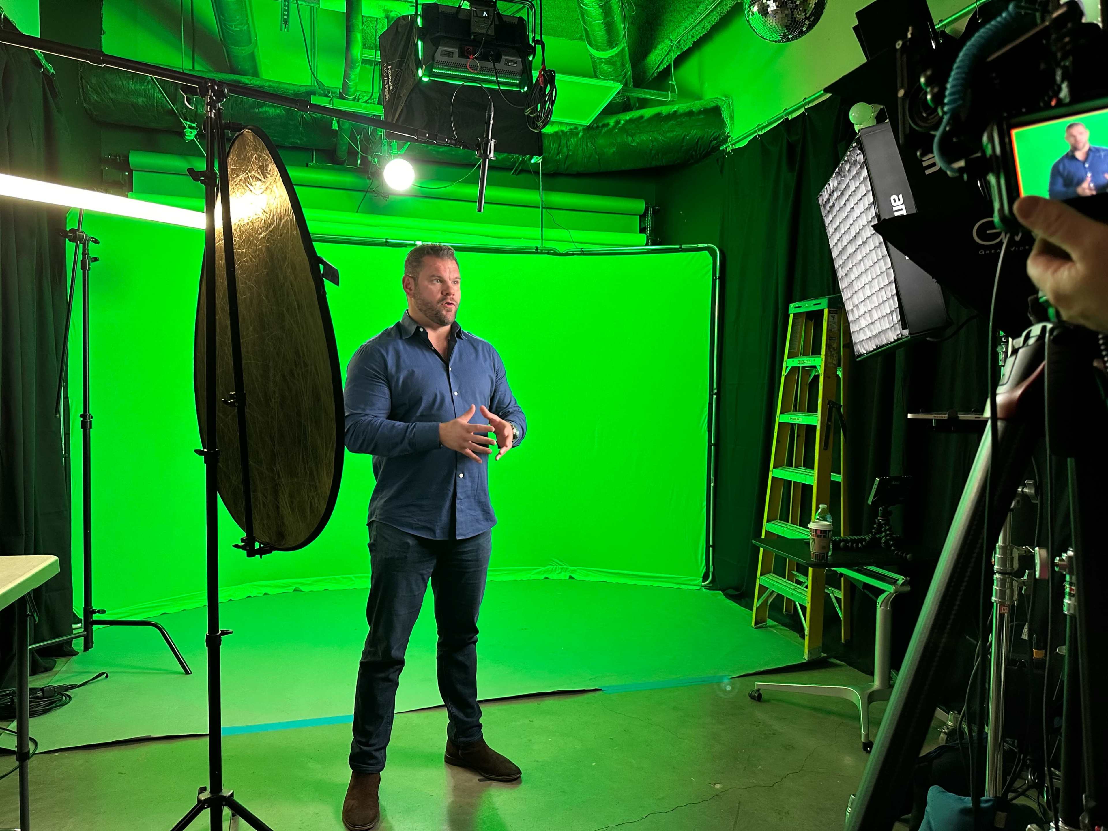 A man stands in front of a green screen, surrounded by various studio lighting equipment and a camera setup.