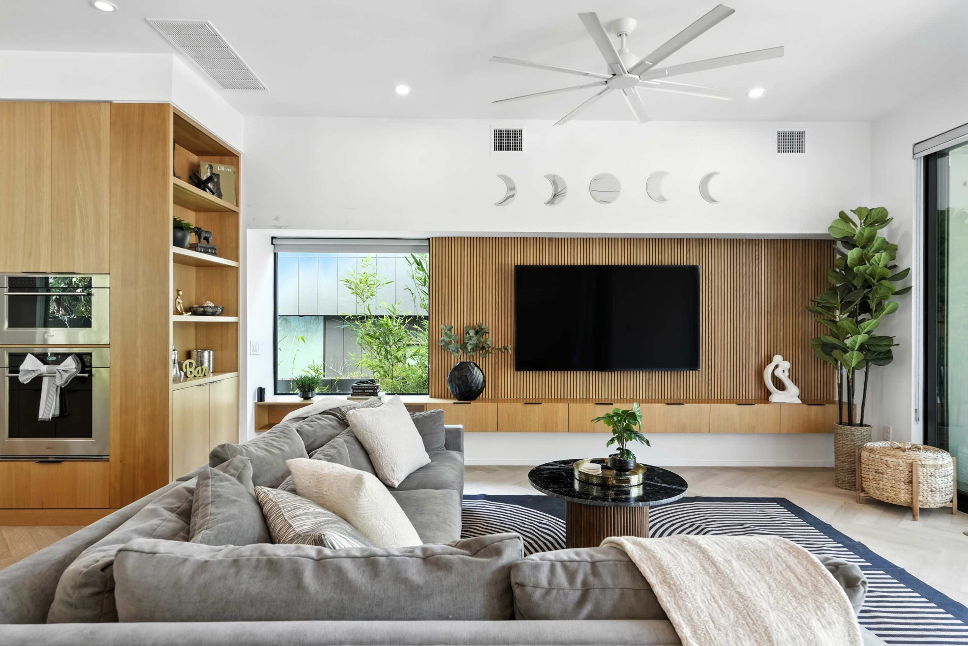 The living room features a large sectional sofa, a coffee table, a wall-mounted television, and decorative elements like plants and artwork on wooden shelving.