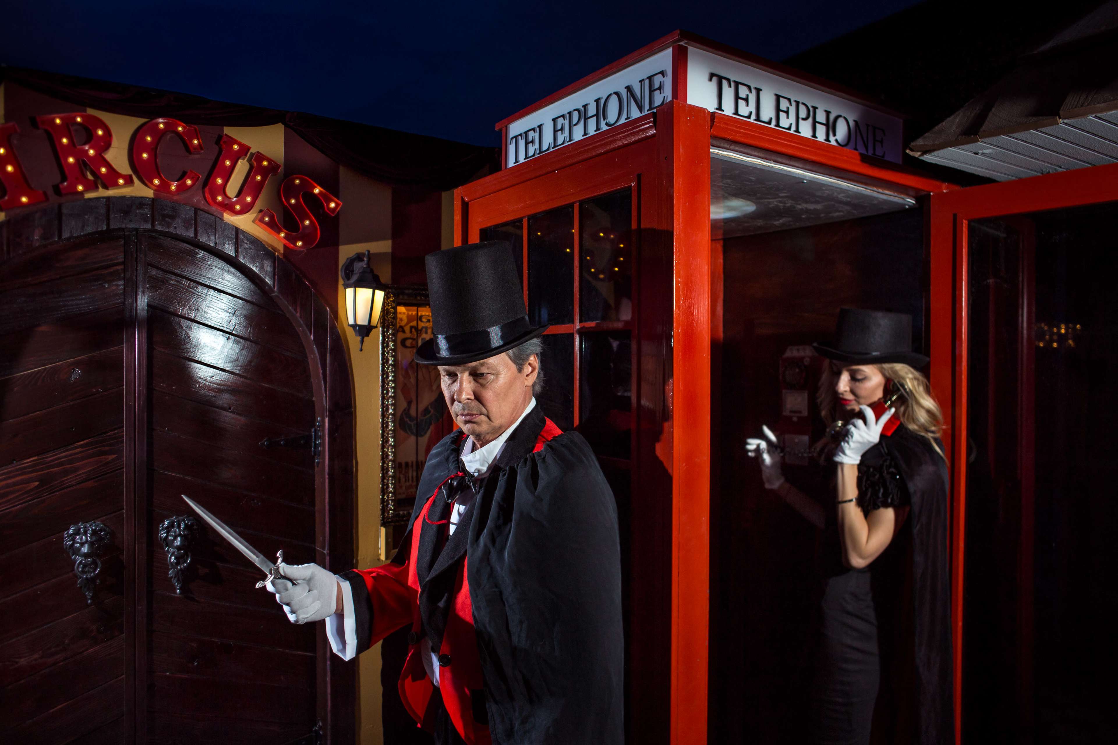 A man in a top hat and red coat stands beside a vintage telephone booth, while a woman in a black outfit stands inside the booth.