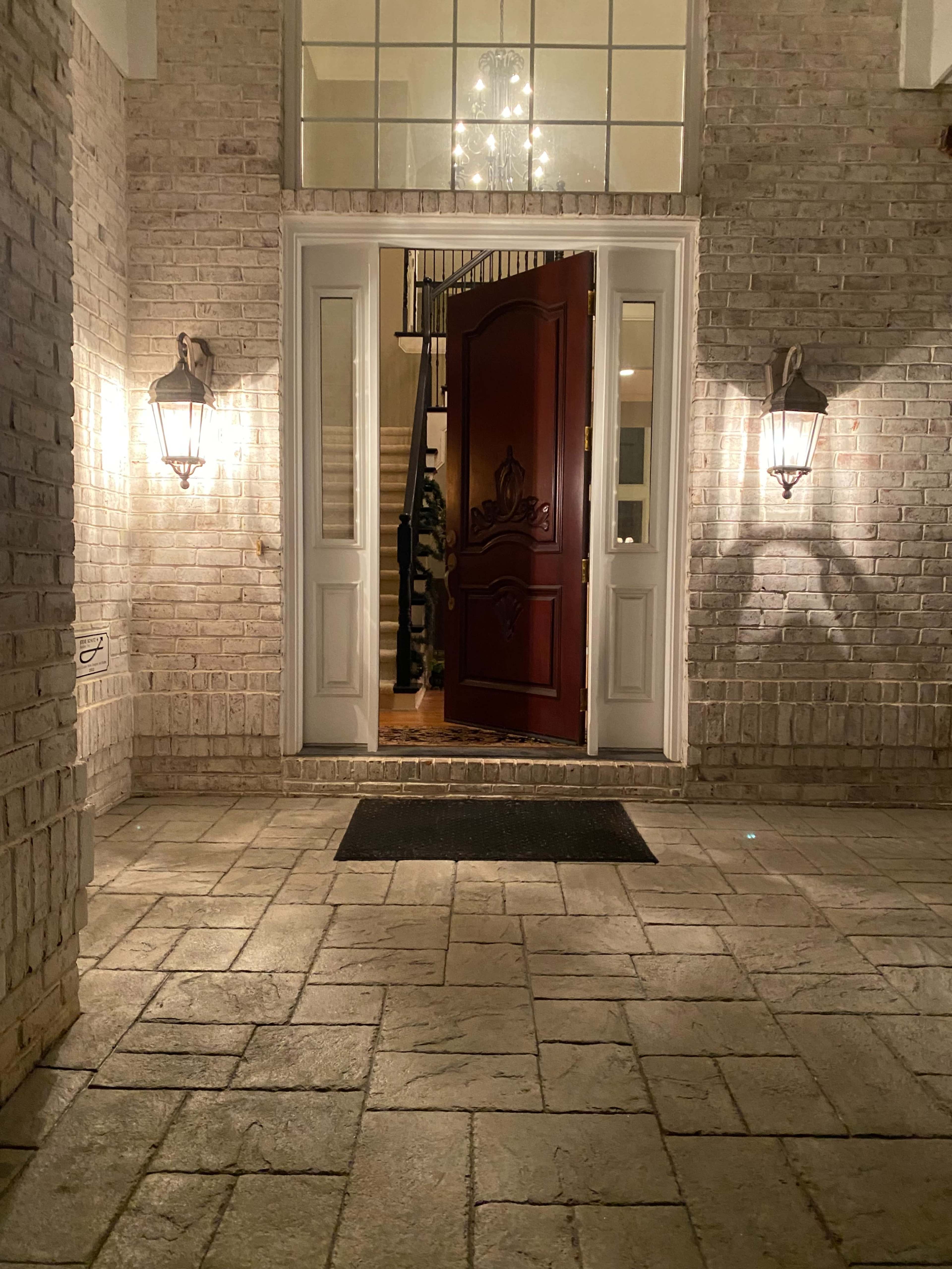 A large front entrance of a house with a red double door slightly open, flanked by two lanterns illuminating the stone pathway leading up to it.