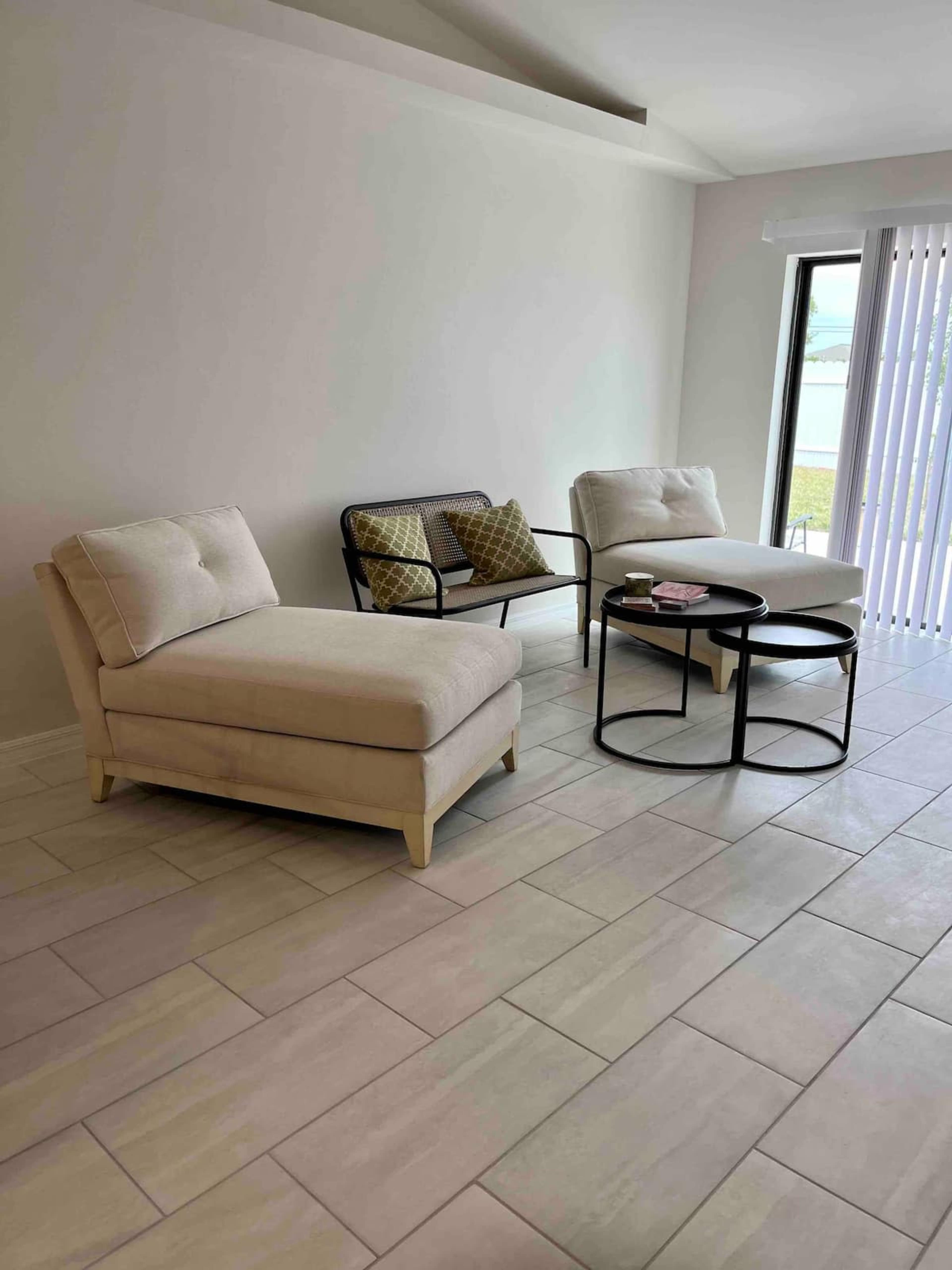 A light-colored living area with two chaise lounges, a small round coffee table, and large windows with vertical blinds.