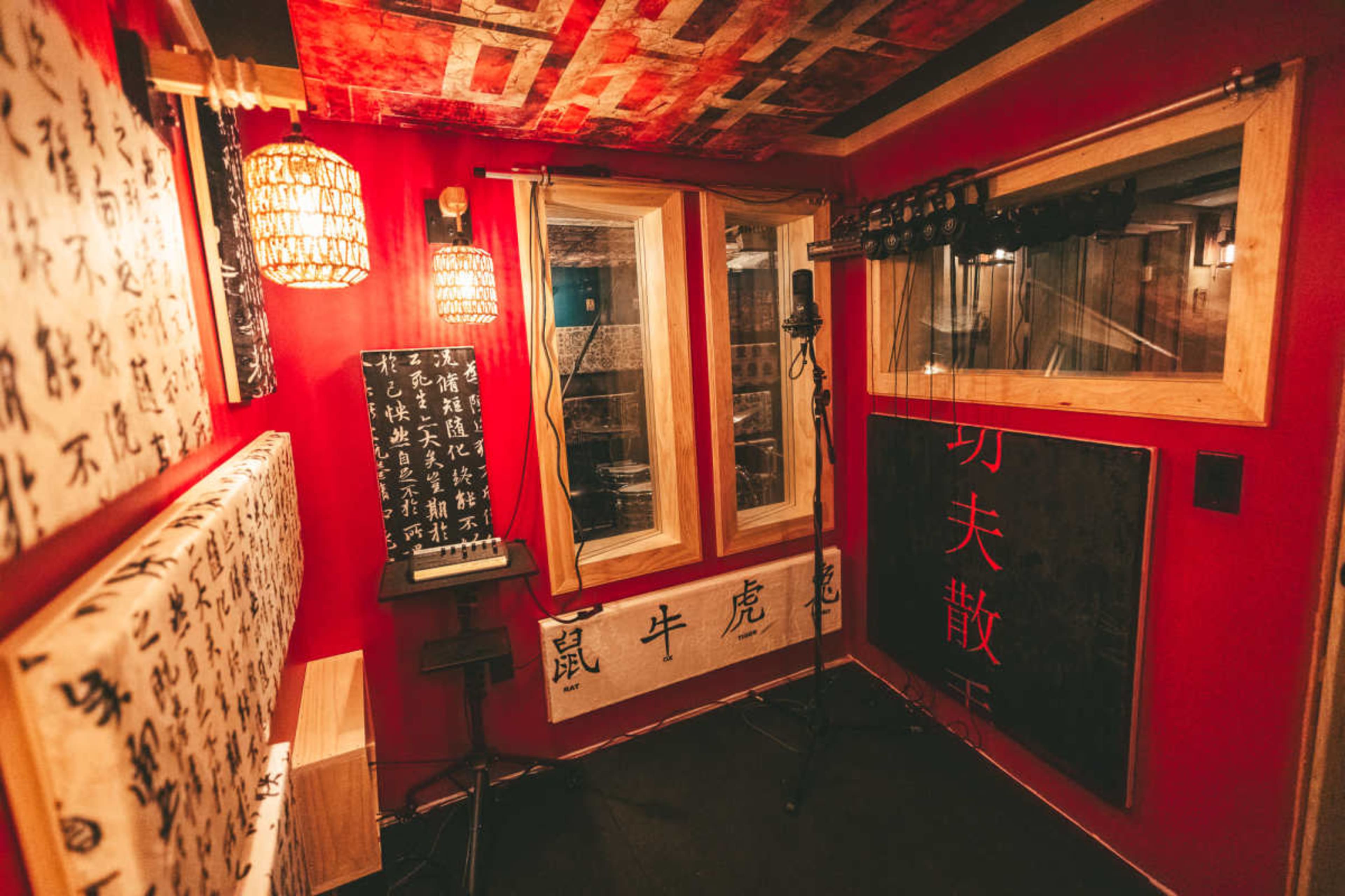 The image shows a soundproof recording studio featuring red walls adorned with various black and white decorative elements and multiple microphones set up for recording.