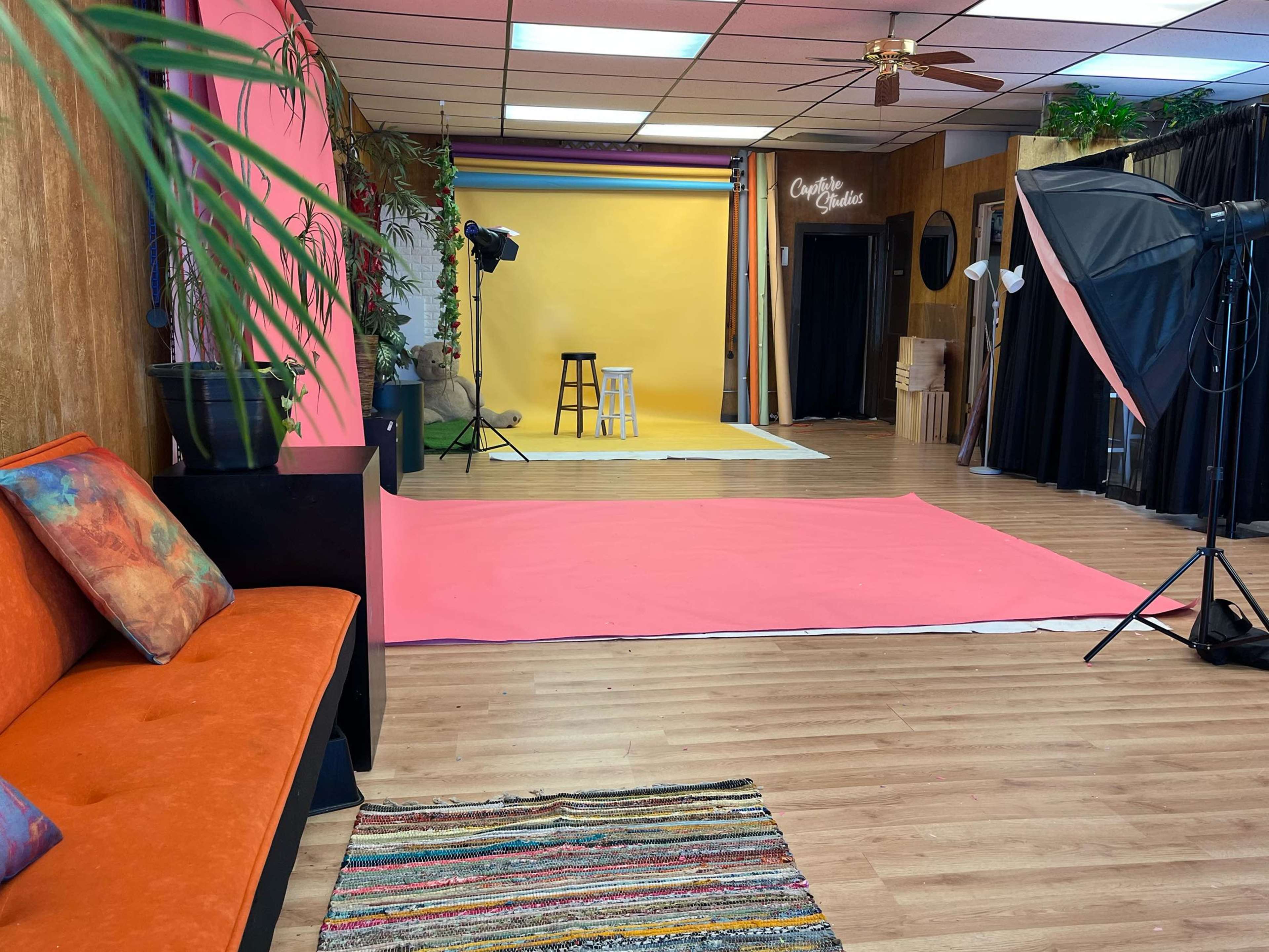 The image shows a brightly colored studio space with pink and yellow backdrops, a stool, and various photography equipment.