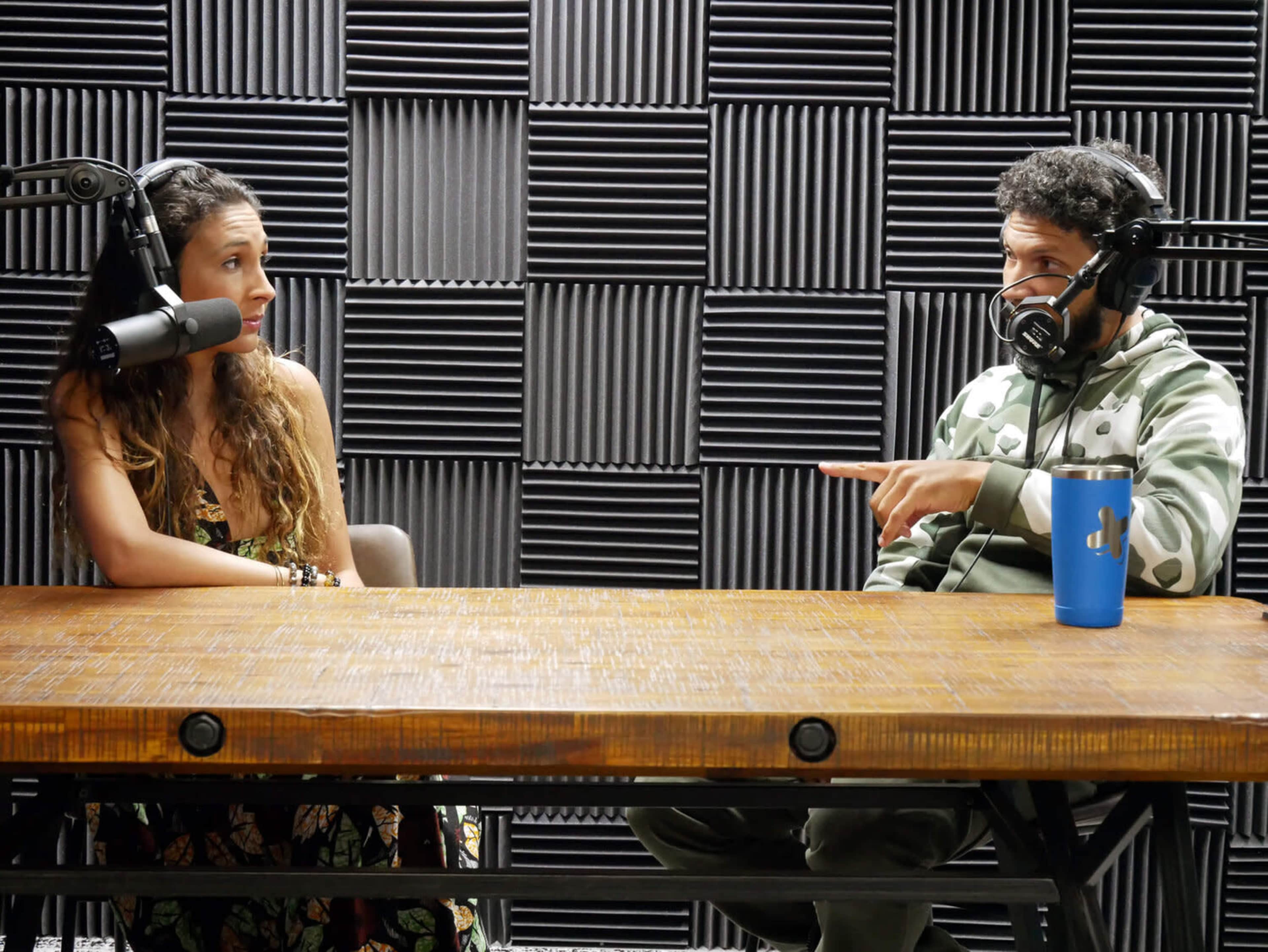 A woman and a man are seated at a wooden table in a recording studio with soundproofing panels, engaging in conversation while wearing microphones.