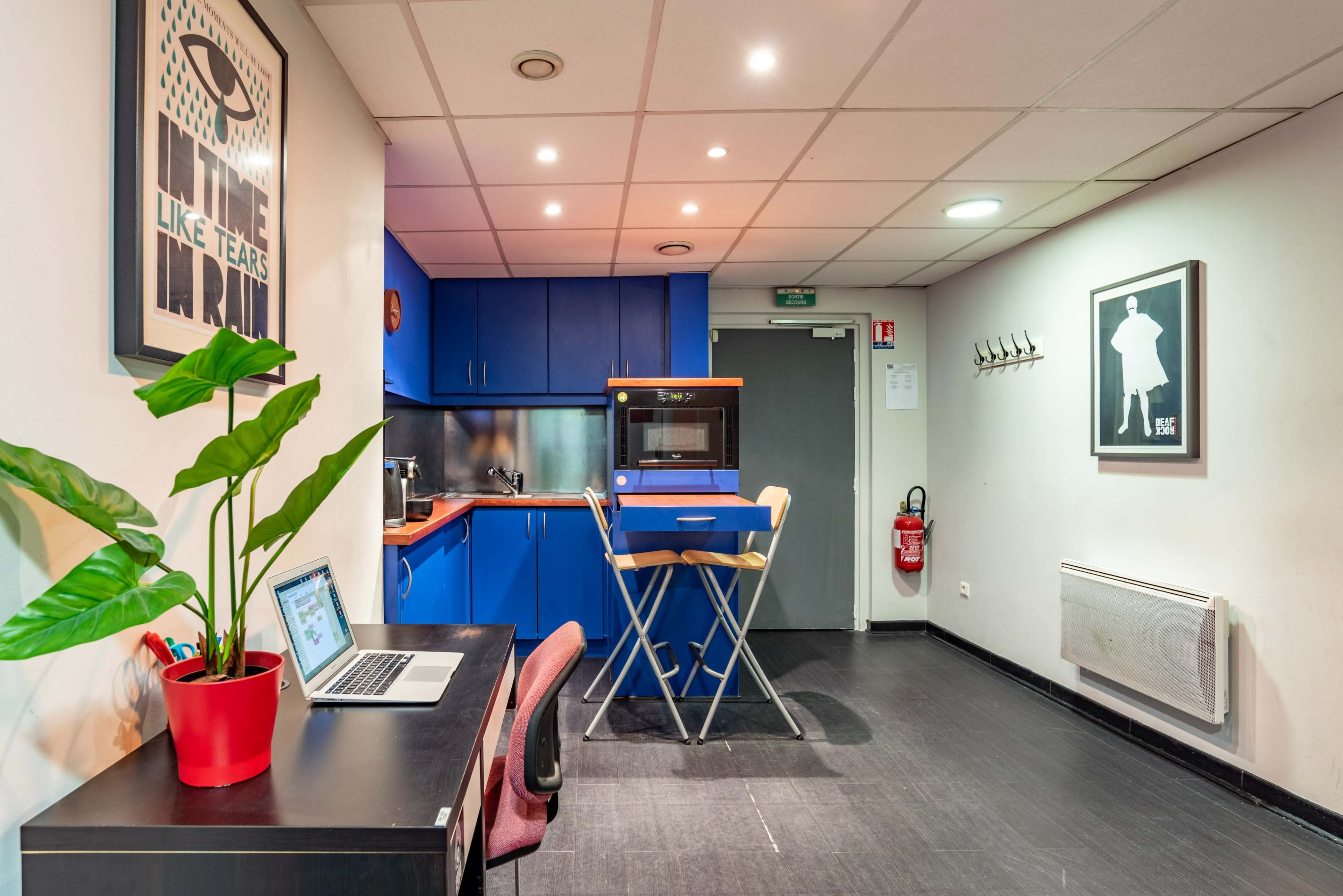 A small kitchen area with blue cabinets, a wooden table with two stools, and a desk with a laptop and a potted plant.