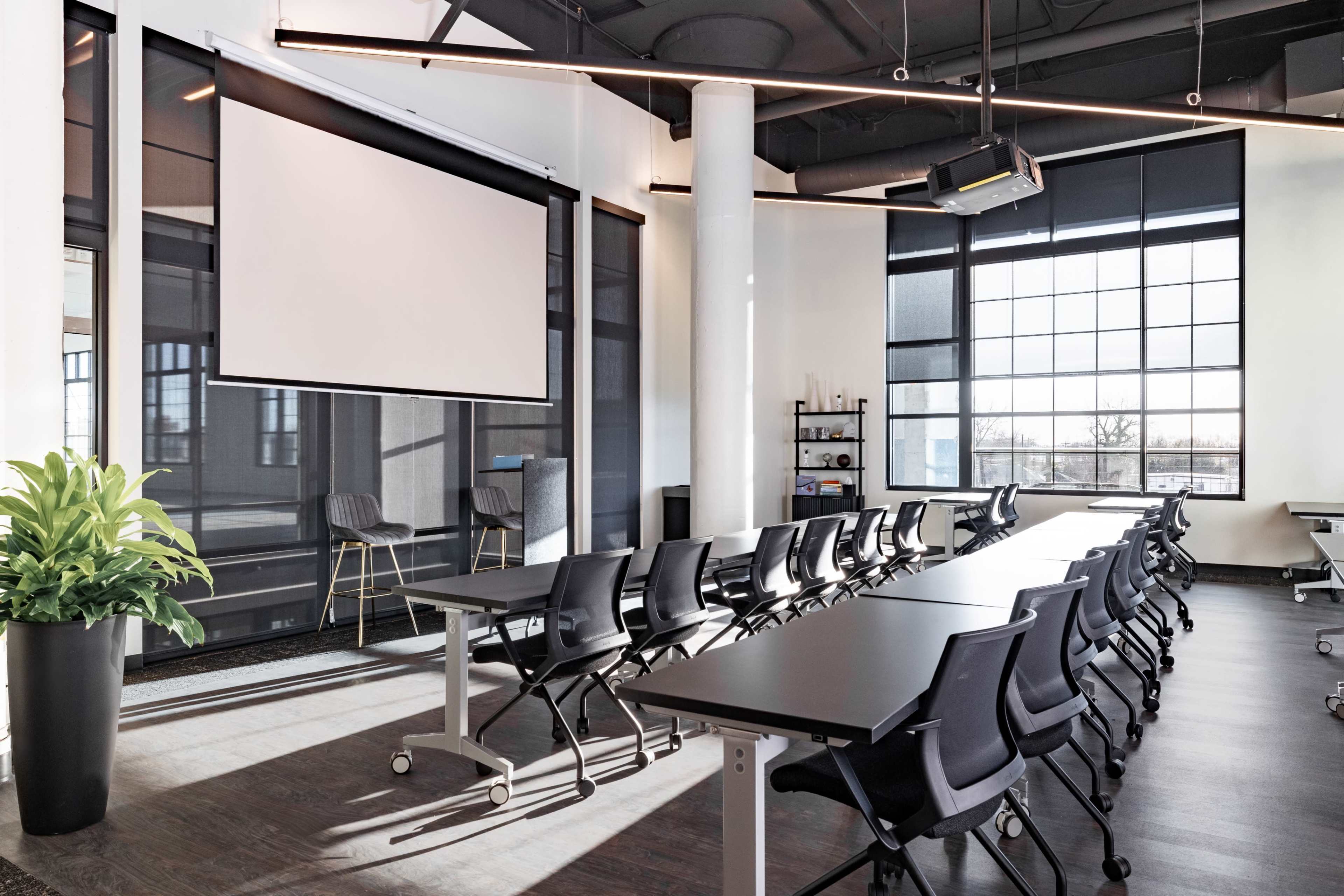 The image shows a modern conference room with tables arranged in a U-shape, a large screen at the front, and large windows providing natural light.