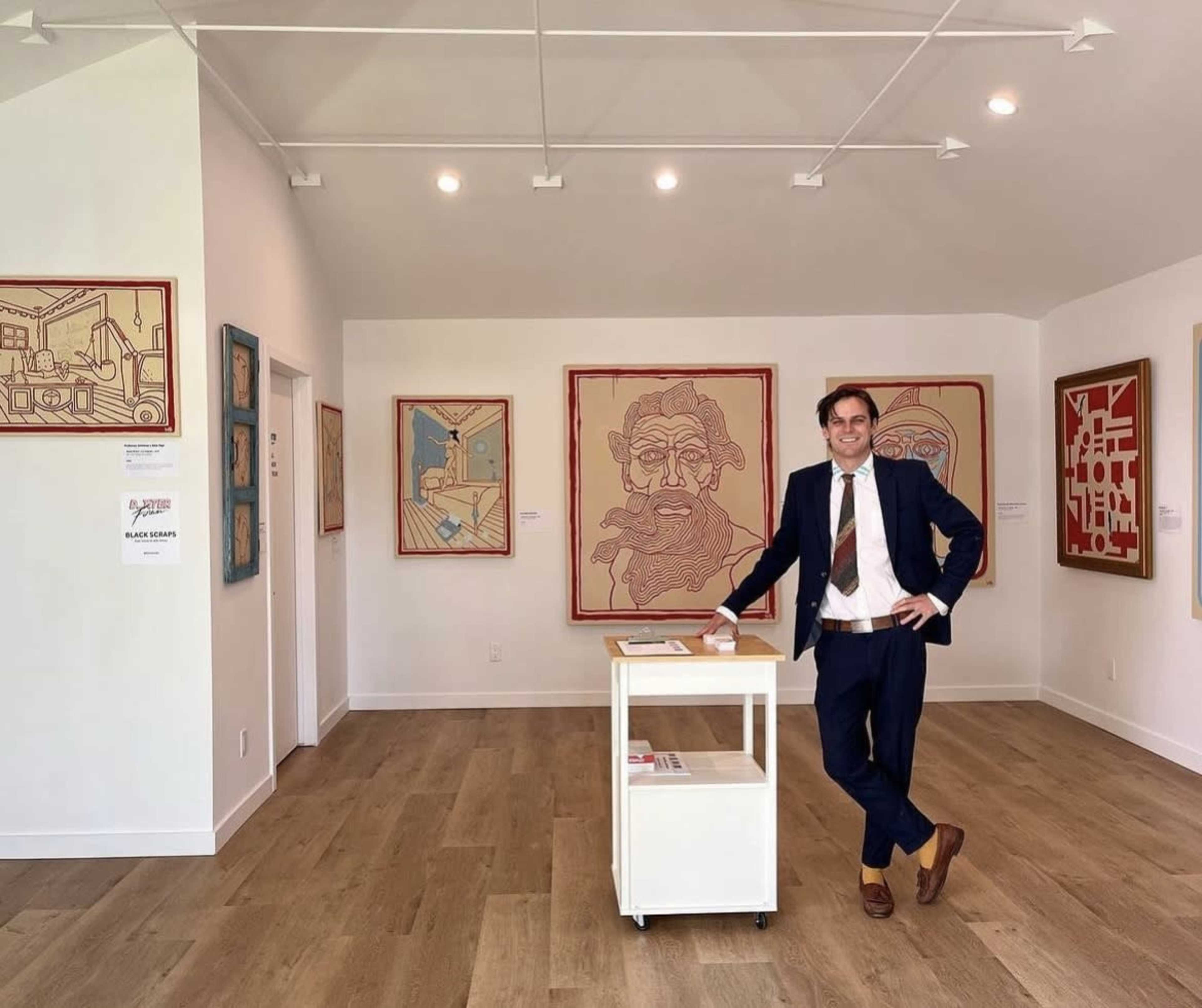 A man in a suit stands beside a display table in an art gallery featuring framed artwork on the walls.