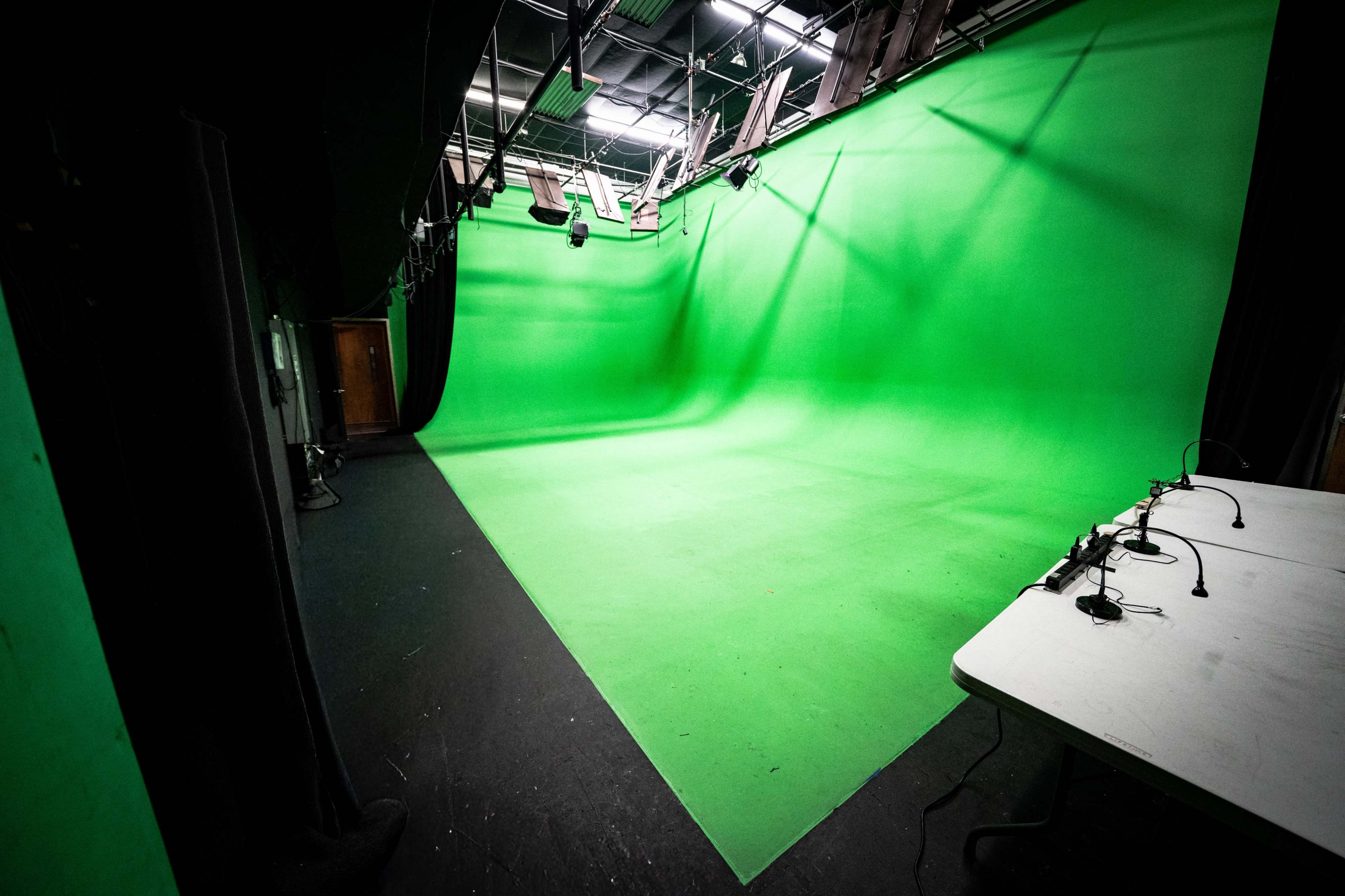 The image shows a large green screen studio with a seamless green background and a table positioned to the side.
