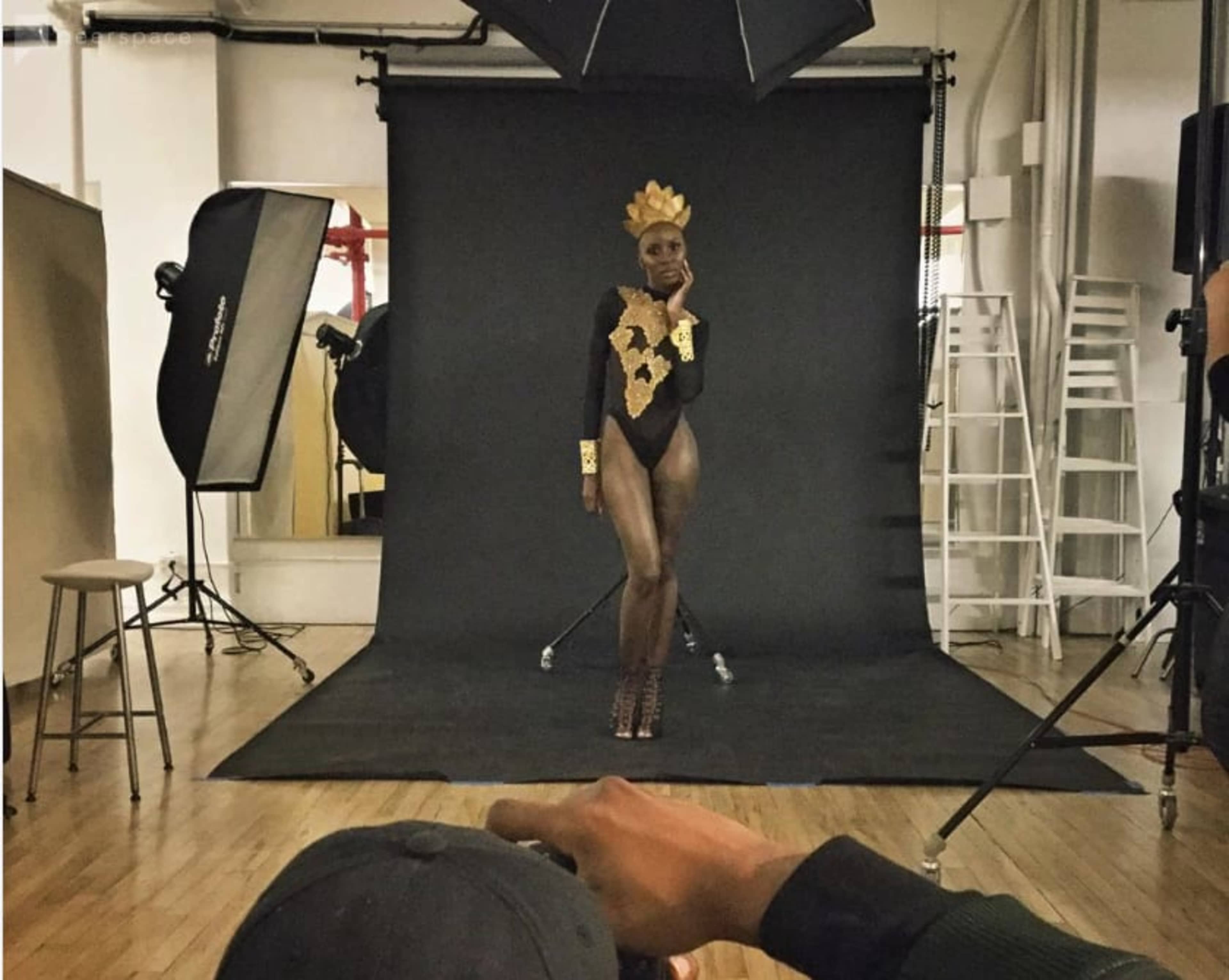 A model poses on a studio set with a black backdrop, surrounded by photography equipment and lighting.