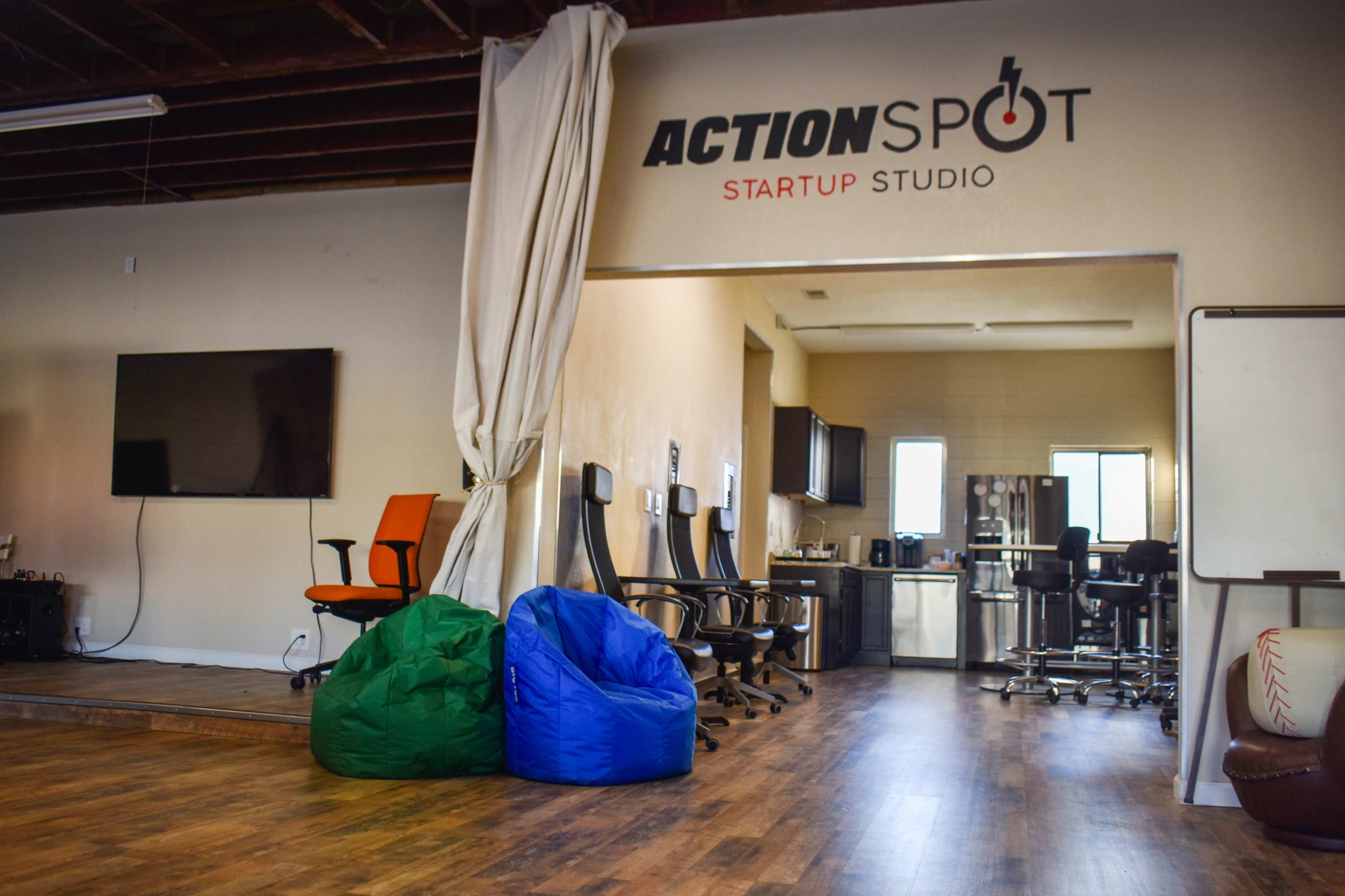 The image shows an open office space featuring colorful bean bags, modern office chairs, and a large wall sign that reads "ACTION SPOT STARTUP STUDIO."
