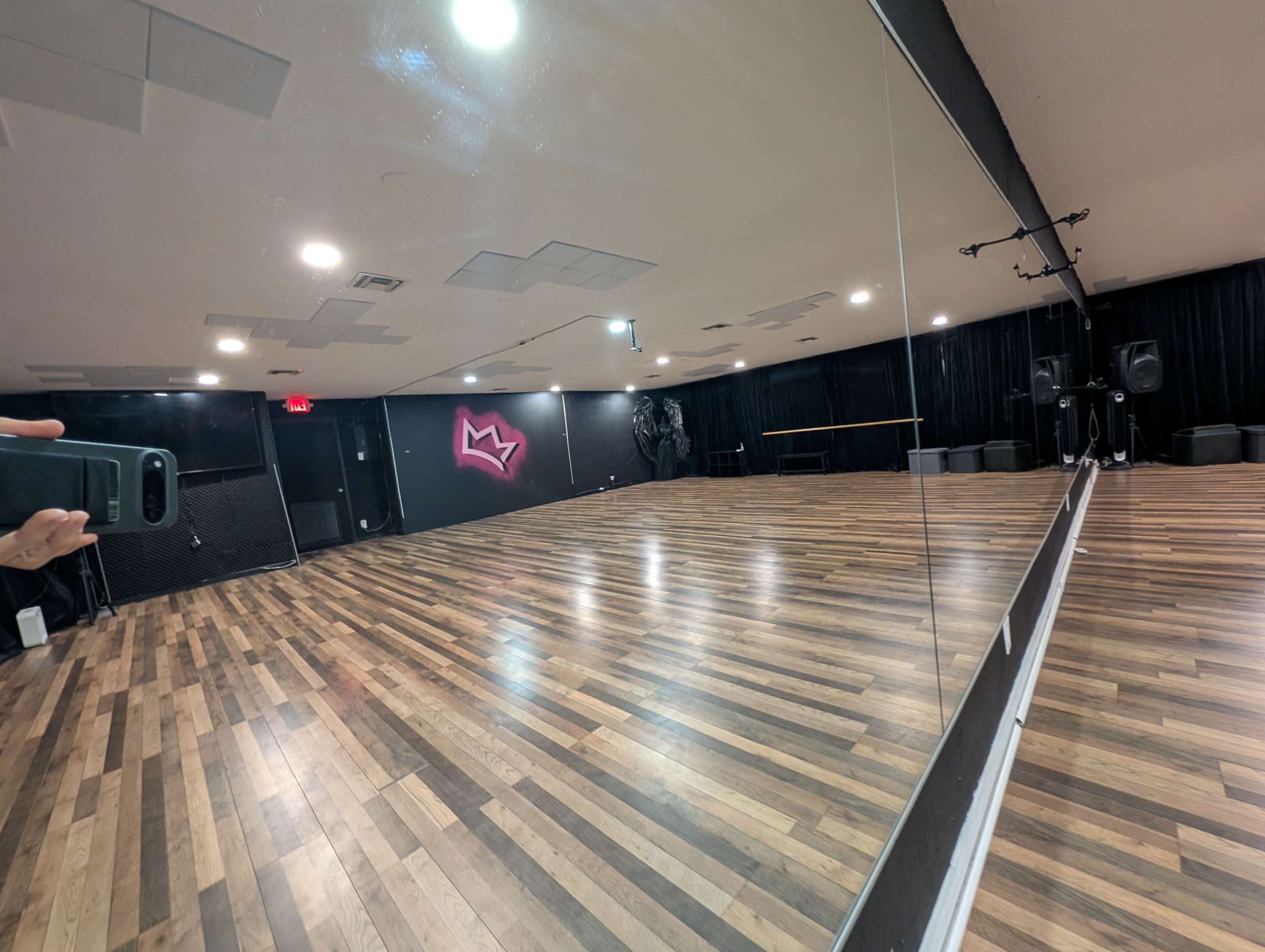 The image shows a dance studio with a wooden floor, a large mirror, and a black wall featuring a pink graphic design.