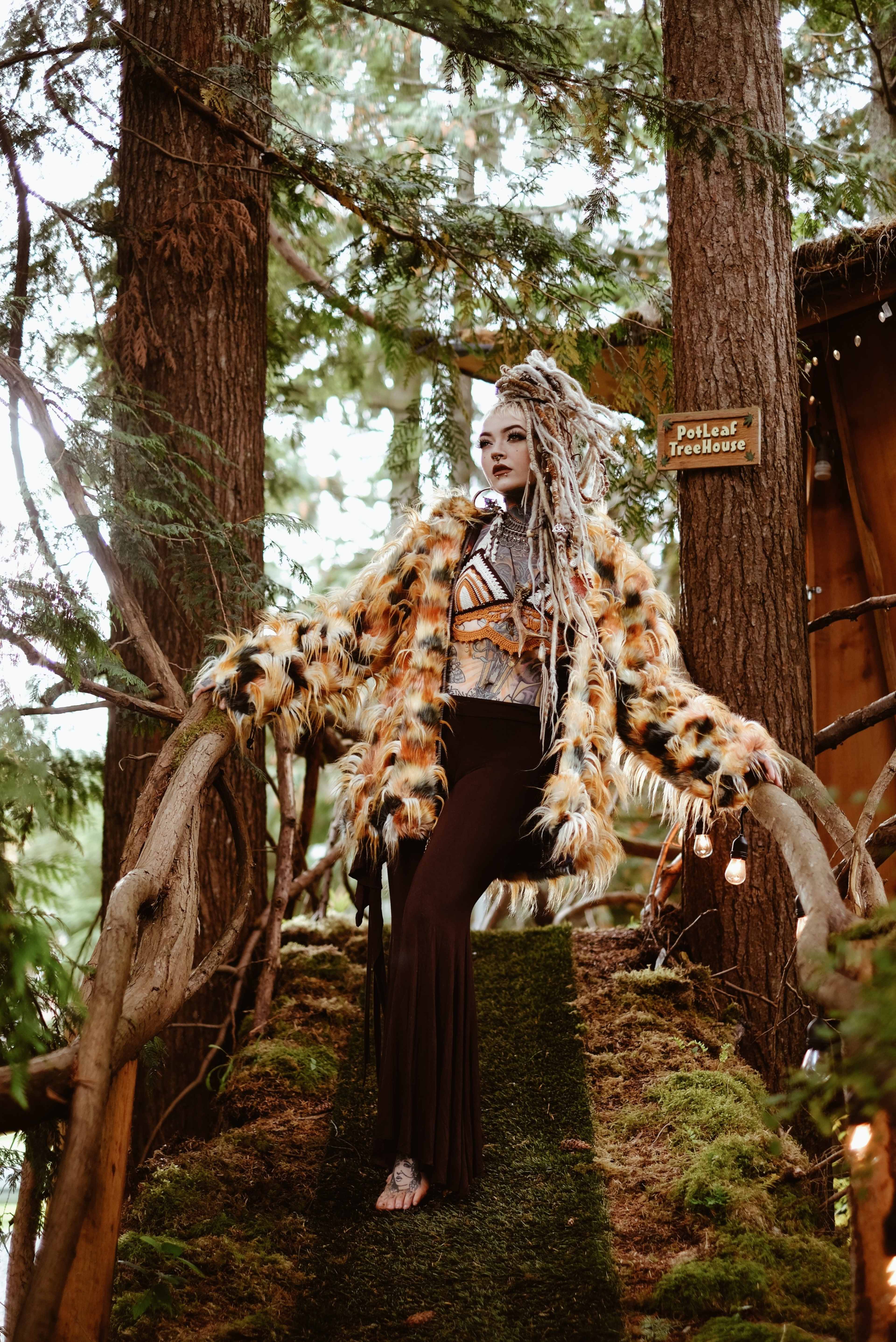 A person with long, decorated hair stands on a wooden bridge surrounded by tall trees, wearing a colorful fur coat and a long skirt.