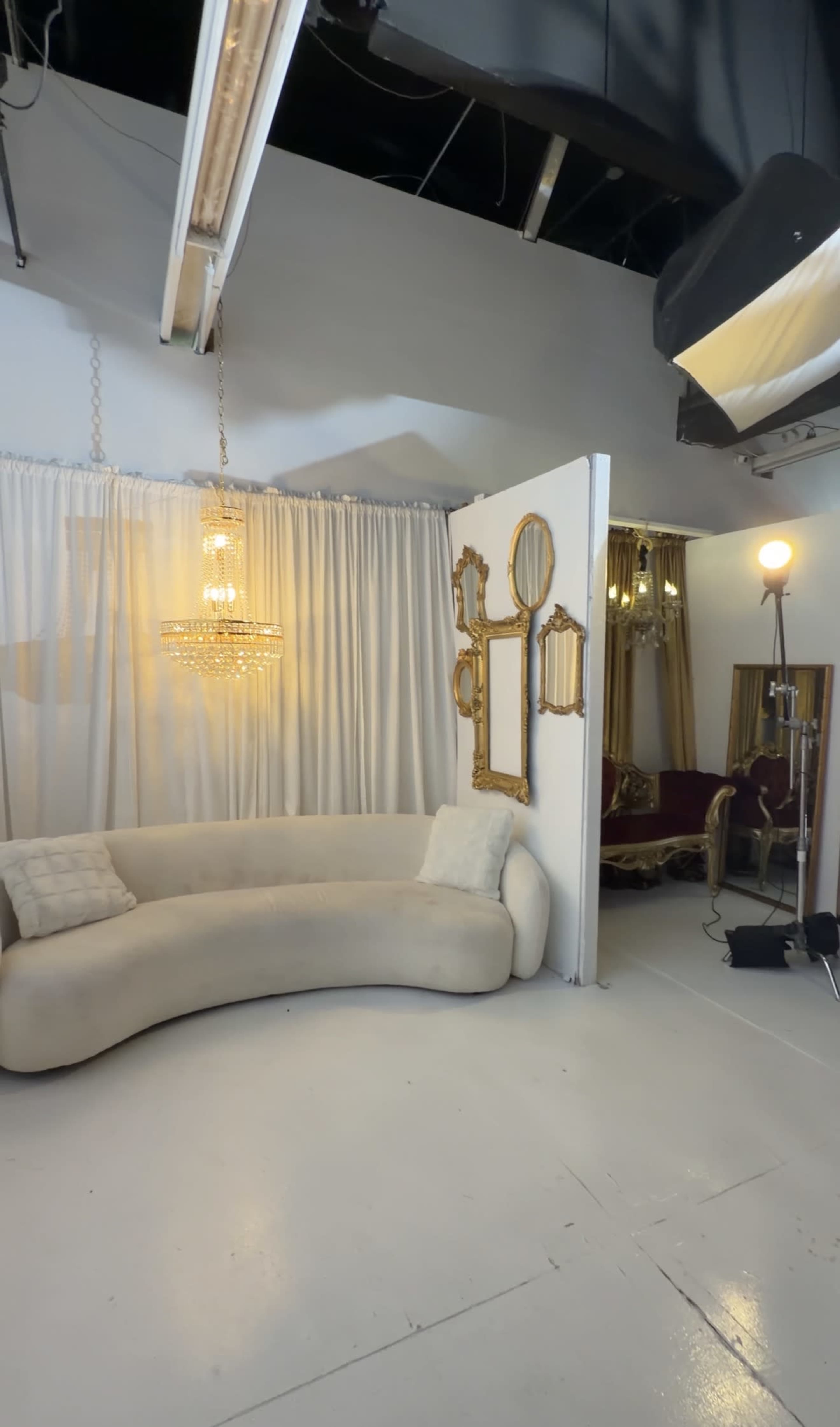 The image shows a studio space with a curved beige sofa, a chandelier, and several decorative mirrors on a partition wall.
