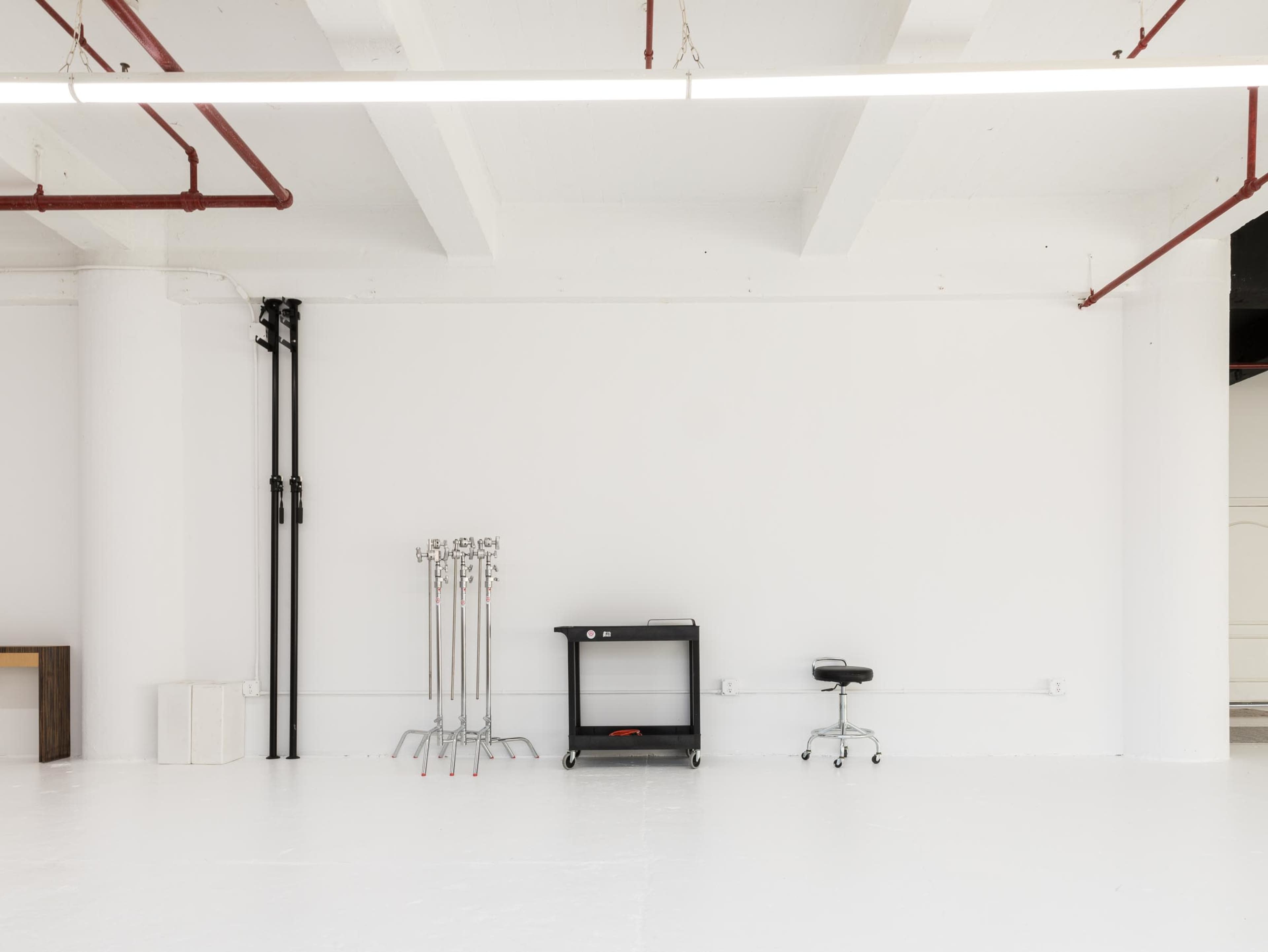 A spacious, empty room with a white floor and walls, featuring a black cart on wheels and a stool against one wall, along with visible pipes.