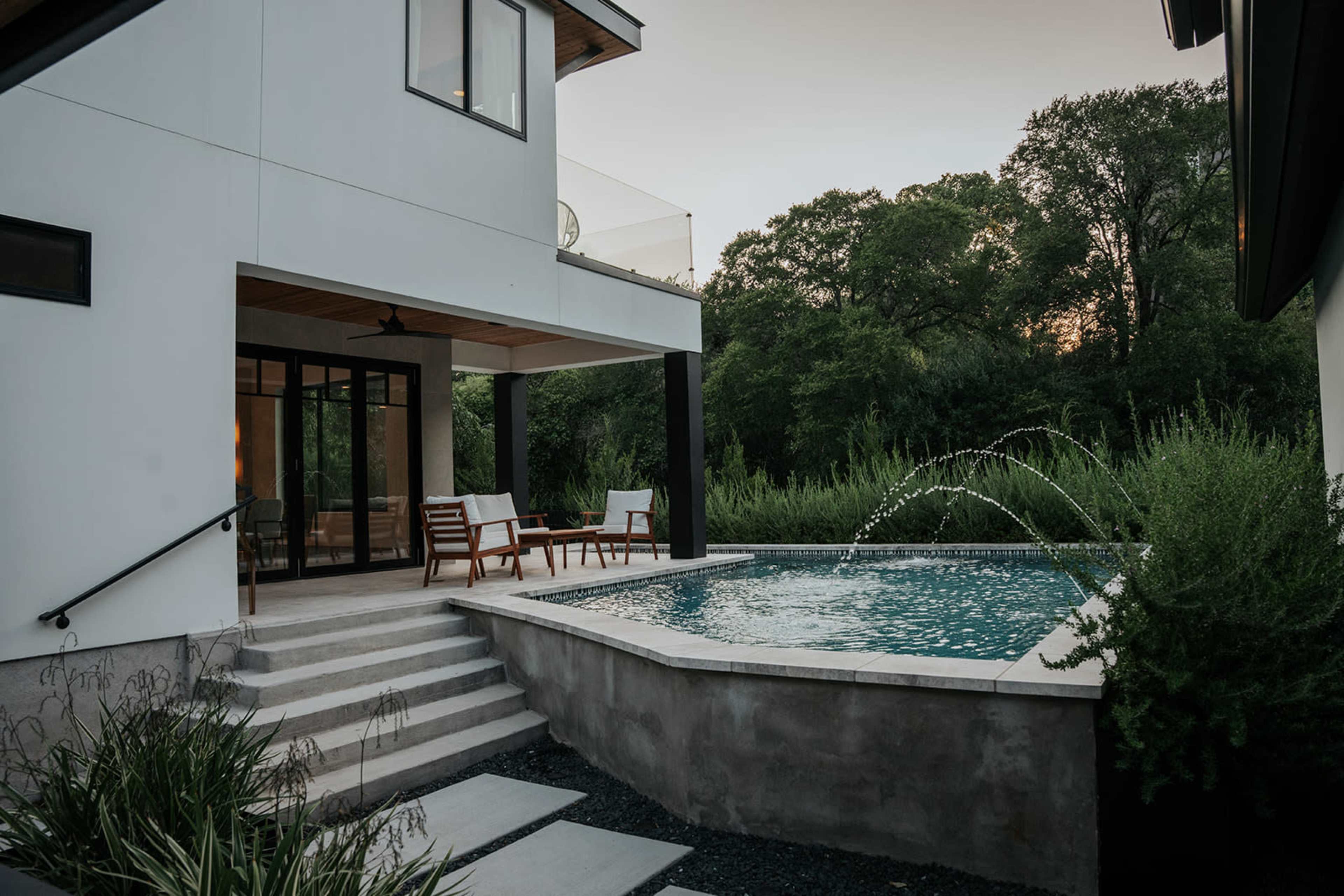 A modern house features a patio with seating and a small pool equipped with a water fountain surrounded by lush greenery.