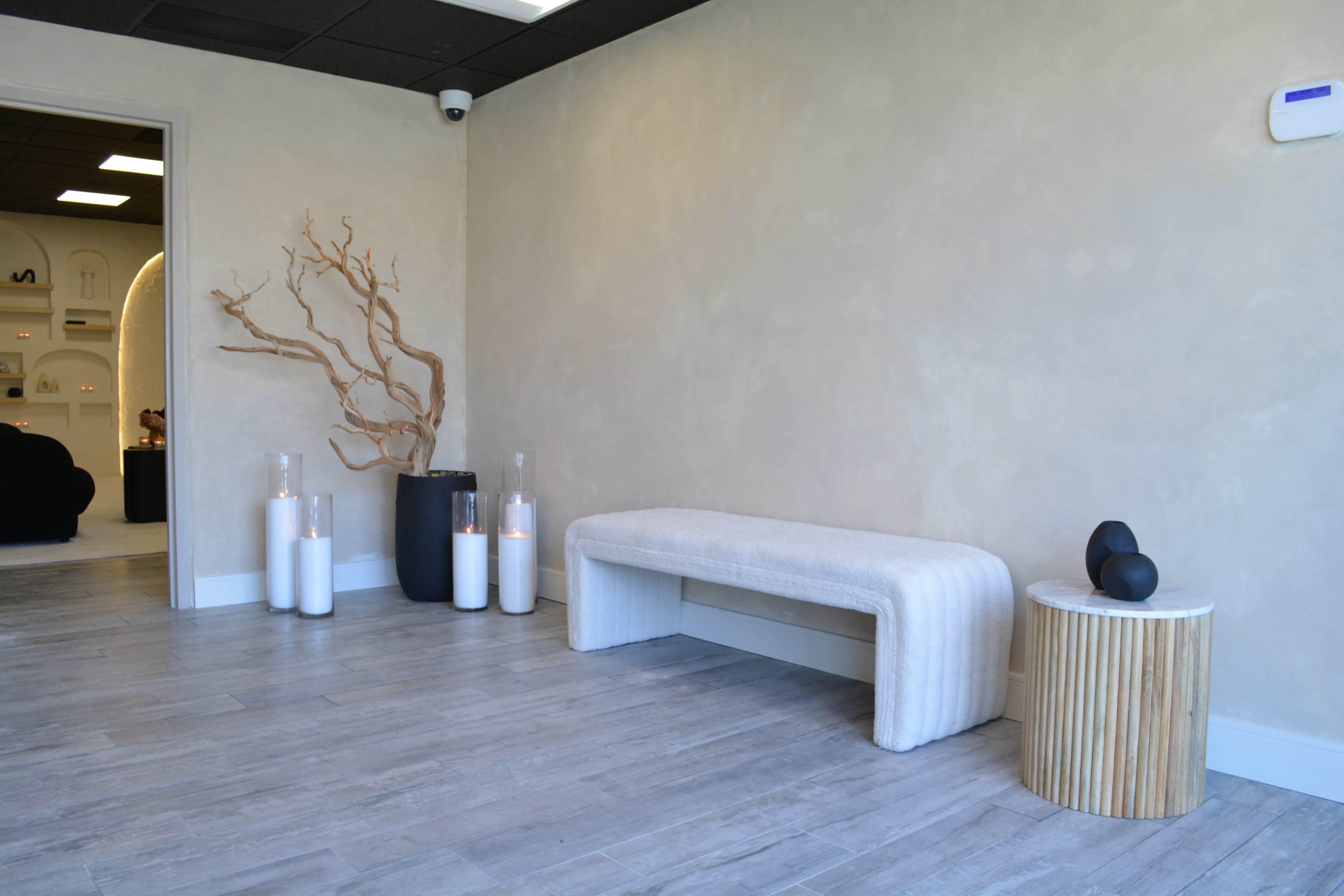 A minimalist interior with a light-colored wall, a textured bench, a wooden side table, and decorative vases on a wooden floor.