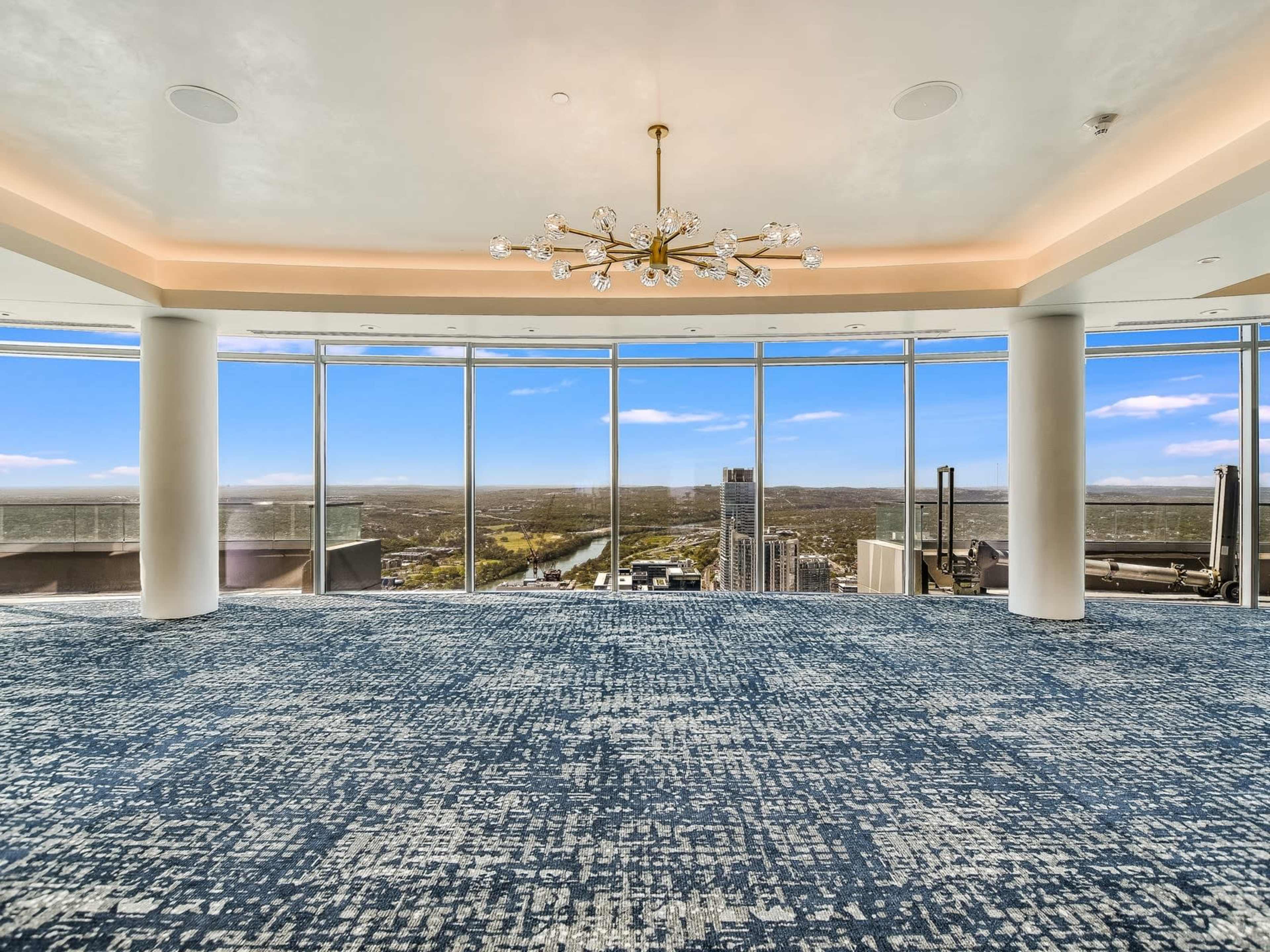 A spacious room with a blue carpet, large windows offering a panoramic view of the landscape, and a chandelier hanging from the ceiling.