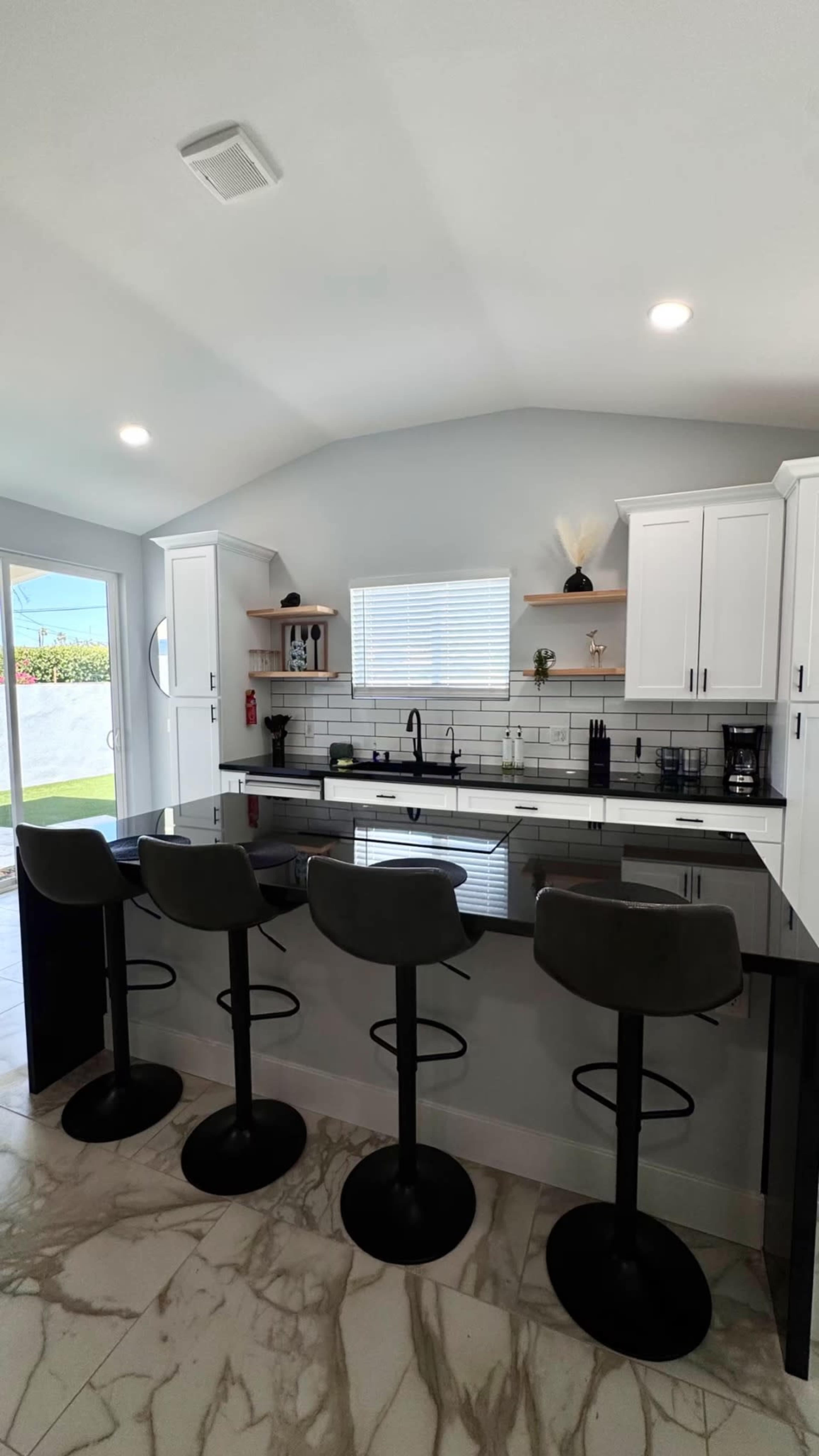 The image shows a modern kitchen with a large island featuring four round bar stools, white cabinets, and a marble-patterned floor.