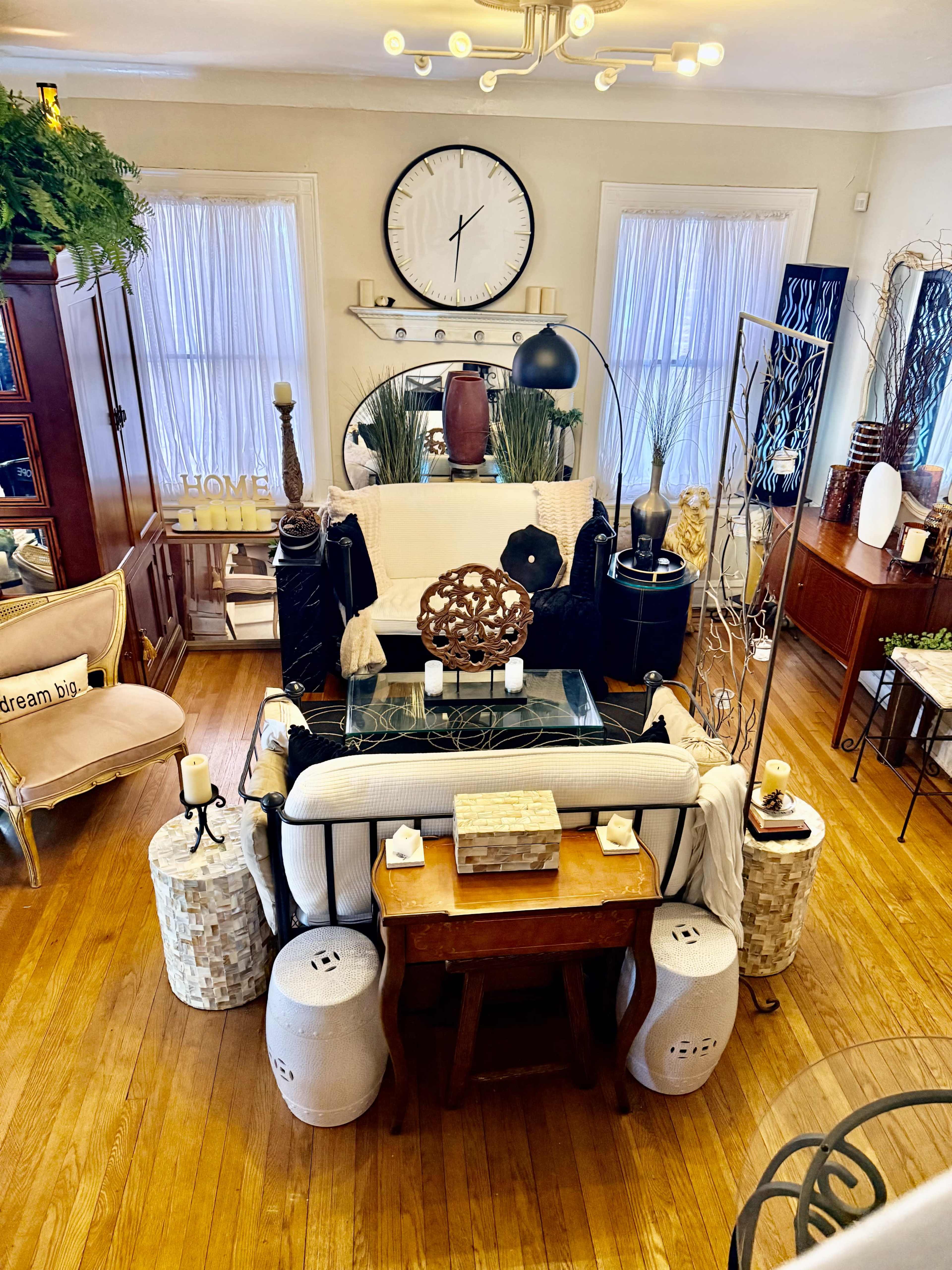 The image shows a cozy living room with a mix of decorative elements, including a large clock, plants, and various furniture pieces arranged around a central seating area.