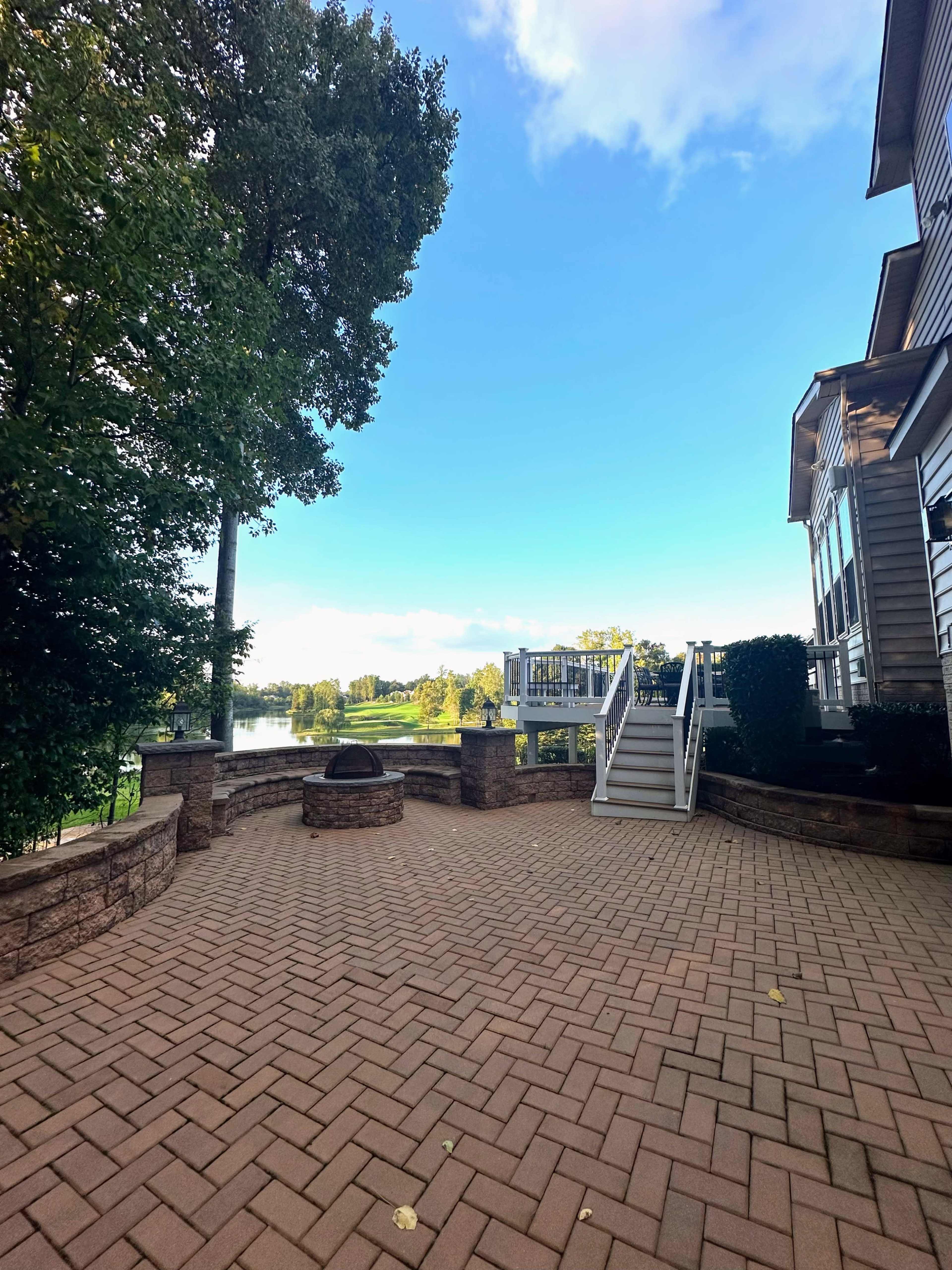 A brick patio with a fire pit overlooks a calm body of water, framed by trees and a house.