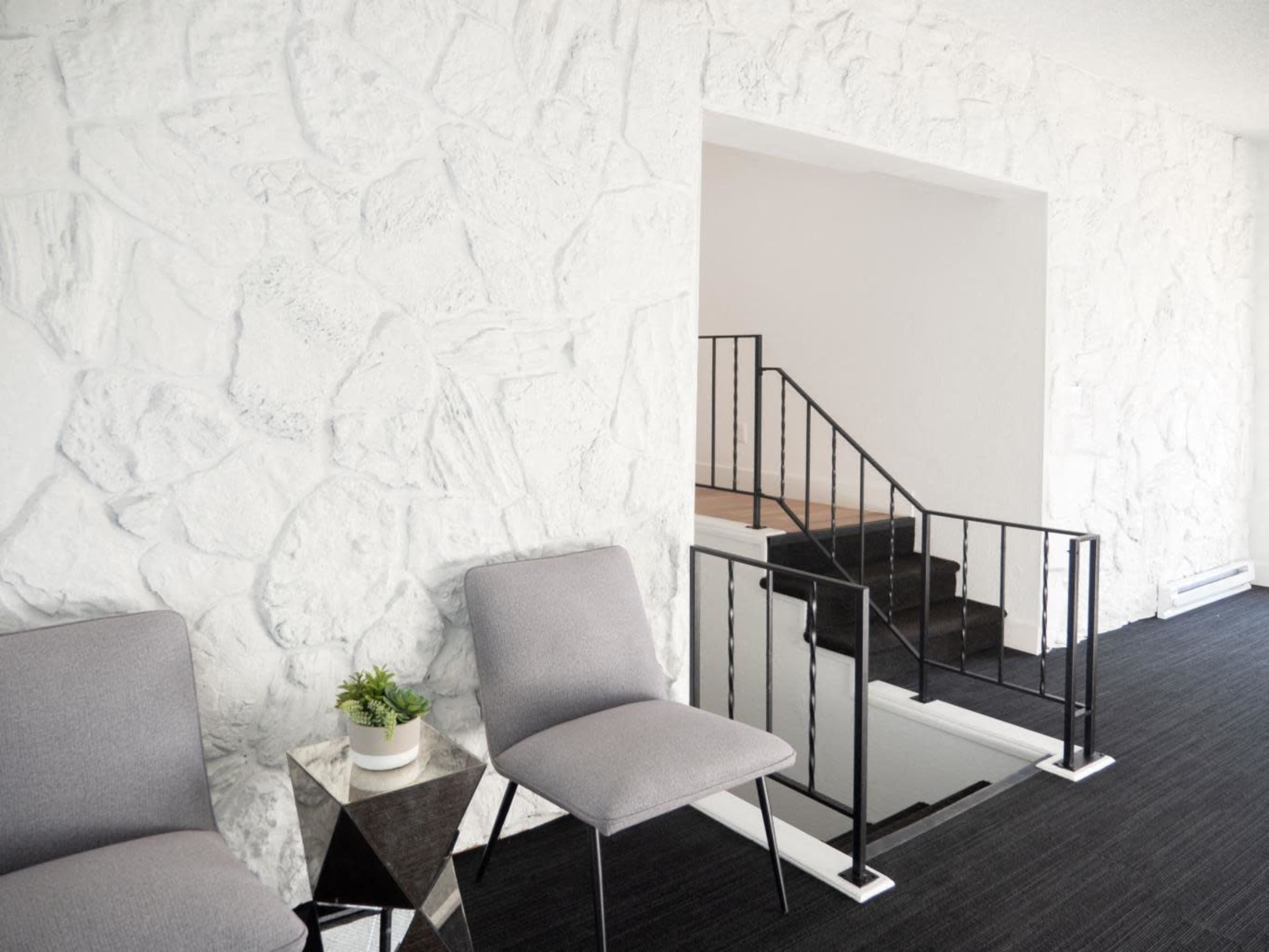 The image shows a minimalist interior with two gray chairs next to a small table, a coffee table, and a staircase leading to a higher level, all against a textured white stone wall.