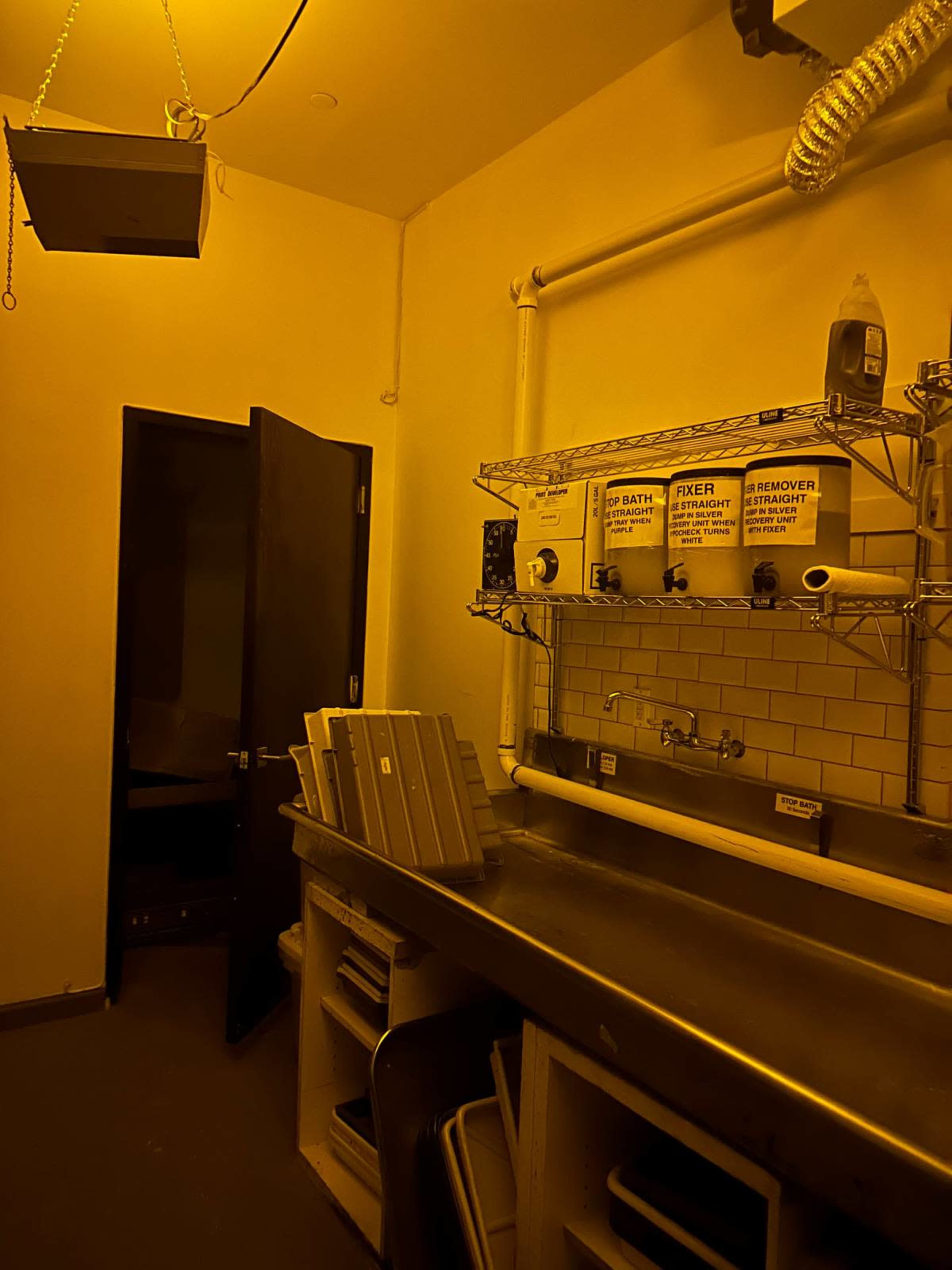 The image depicts a small, dimly-lit room featuring a stainless steel countertop with various cleaning supplies and a door leading to another area.