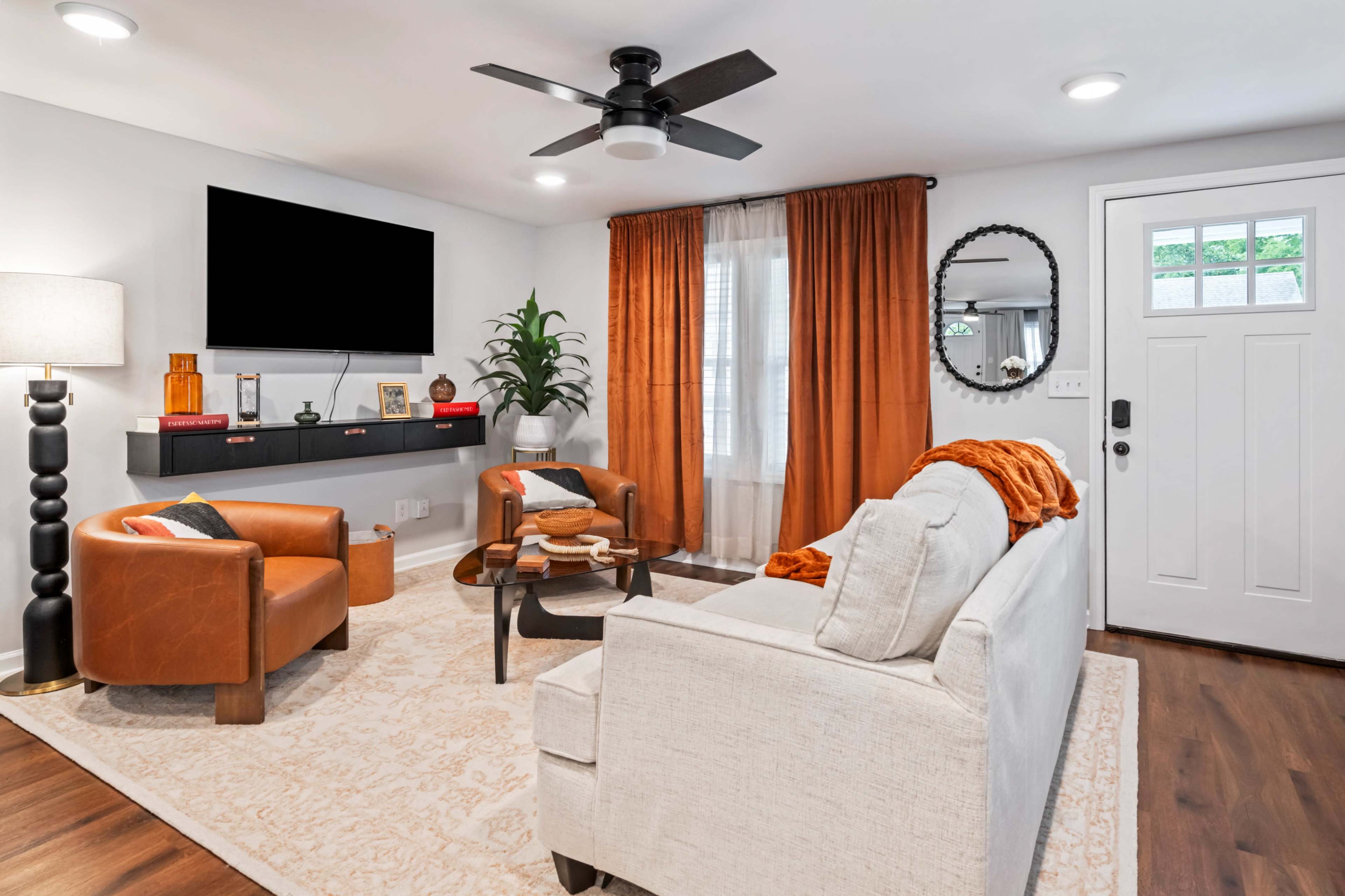 The living room features a neutral sofa, two orange armchairs, a round coffee table, and a wall-mounted television, illuminated by natural light from a nearby window.