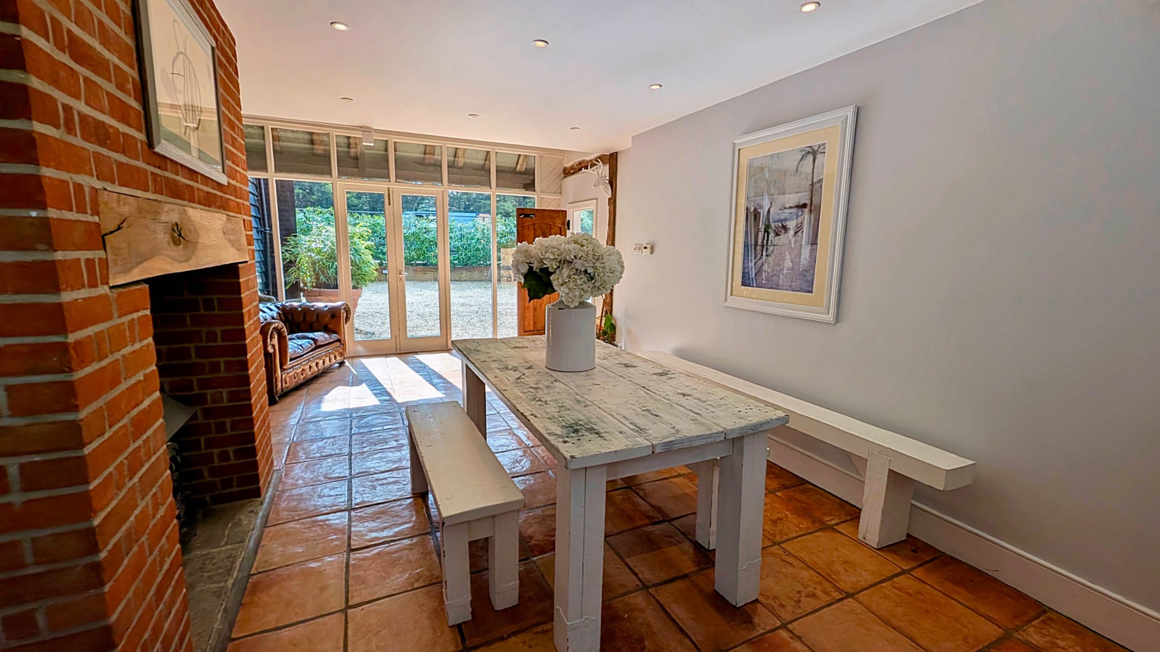 A rustic dining area features a white wooden table and benches, with a brick fireplace and large glass doors leading to a garden.